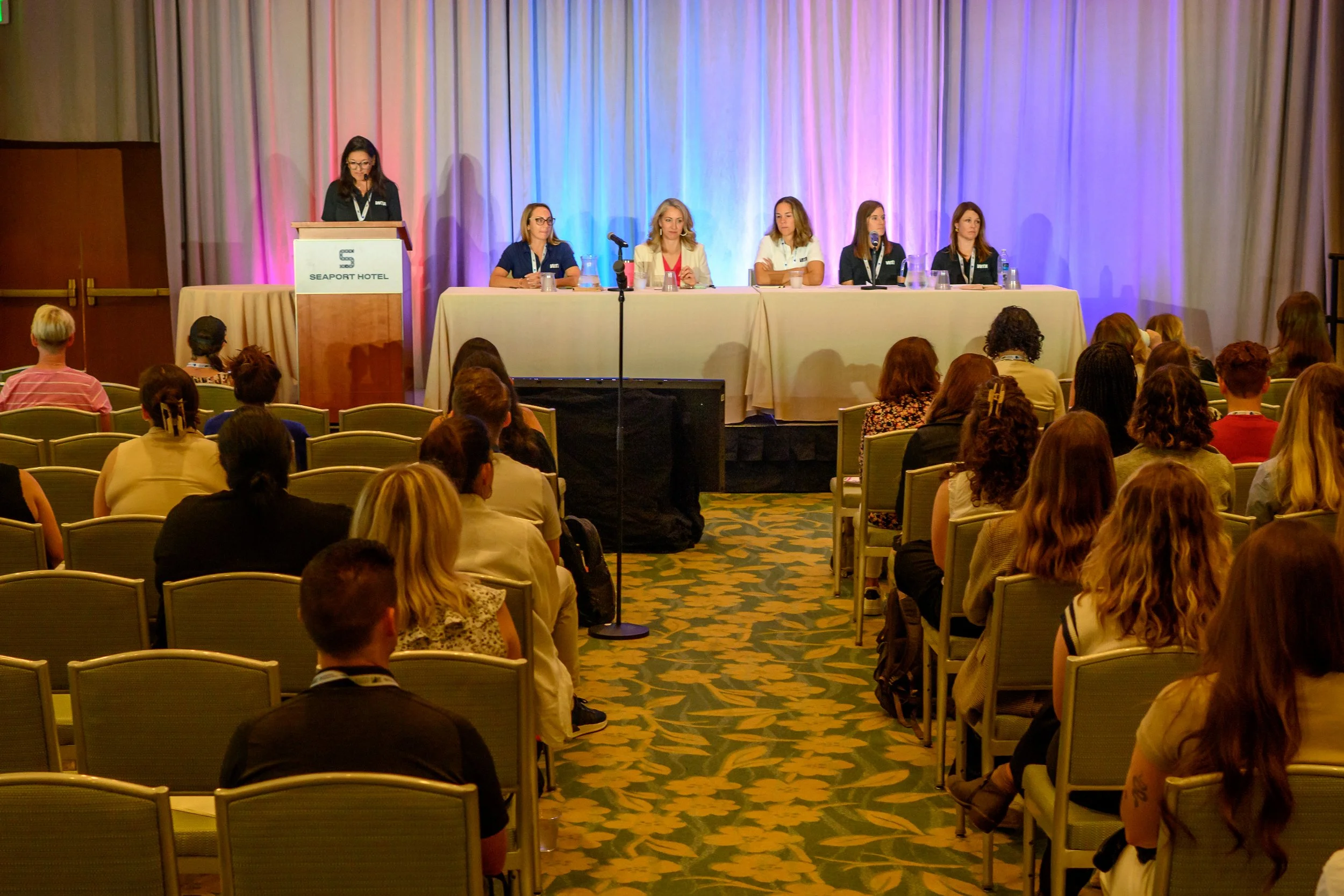 A panel of six women sitting at a long table on a stage in a conference room, with one woman standing at a podium labeled 'Seaport Hotel.' The audience, seated in rows, is watching the presentation. The stage backdrop features blue and purple lighting.