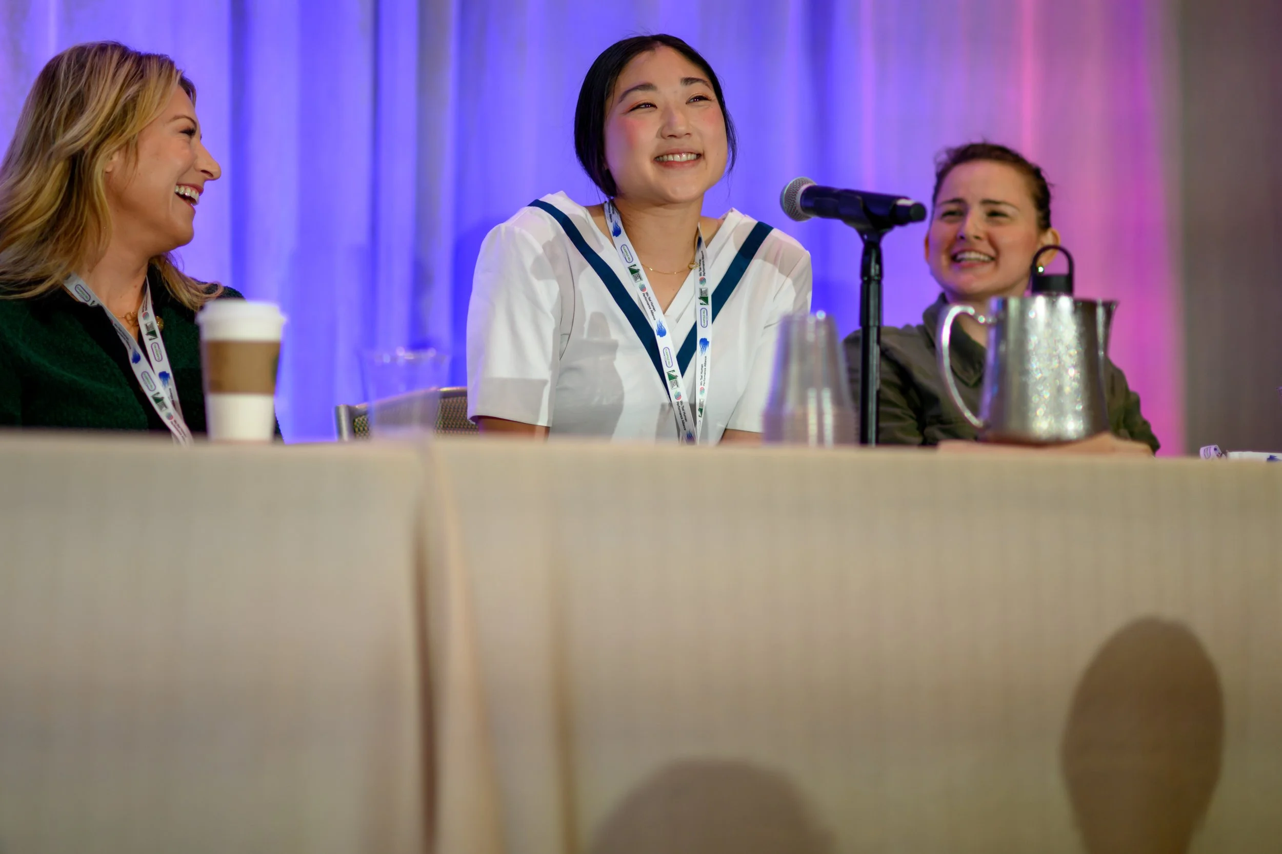 Three women sitting at a conference table, smiling and laughing, with a blue and purple backdrop behind them. The woman in the center has placed her hands on the table, with a microphone in front of her, and is wearing a guitar-shaped lanyard.