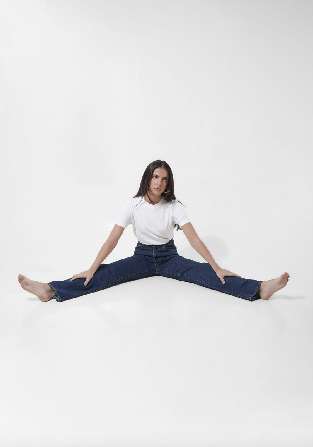Young woman sitting on the floor with legs spread wide apart, hands resting on her legs, looking to the side, wearing a white t-shirt and blue jeans, against a plain white background.