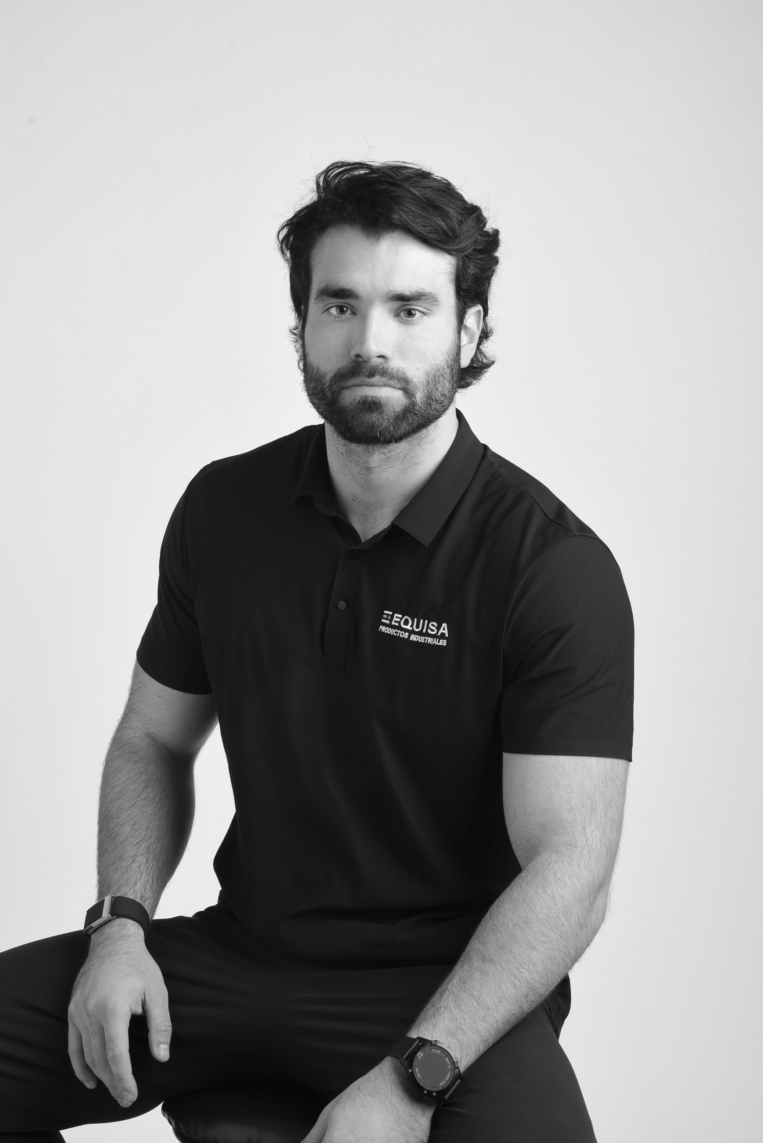 Black and white portrait of a man with dark, wavy hair and a beard, wearing a black polo shirt with a logo. He is sitting with one hand resting on his knee, against a plain light background.