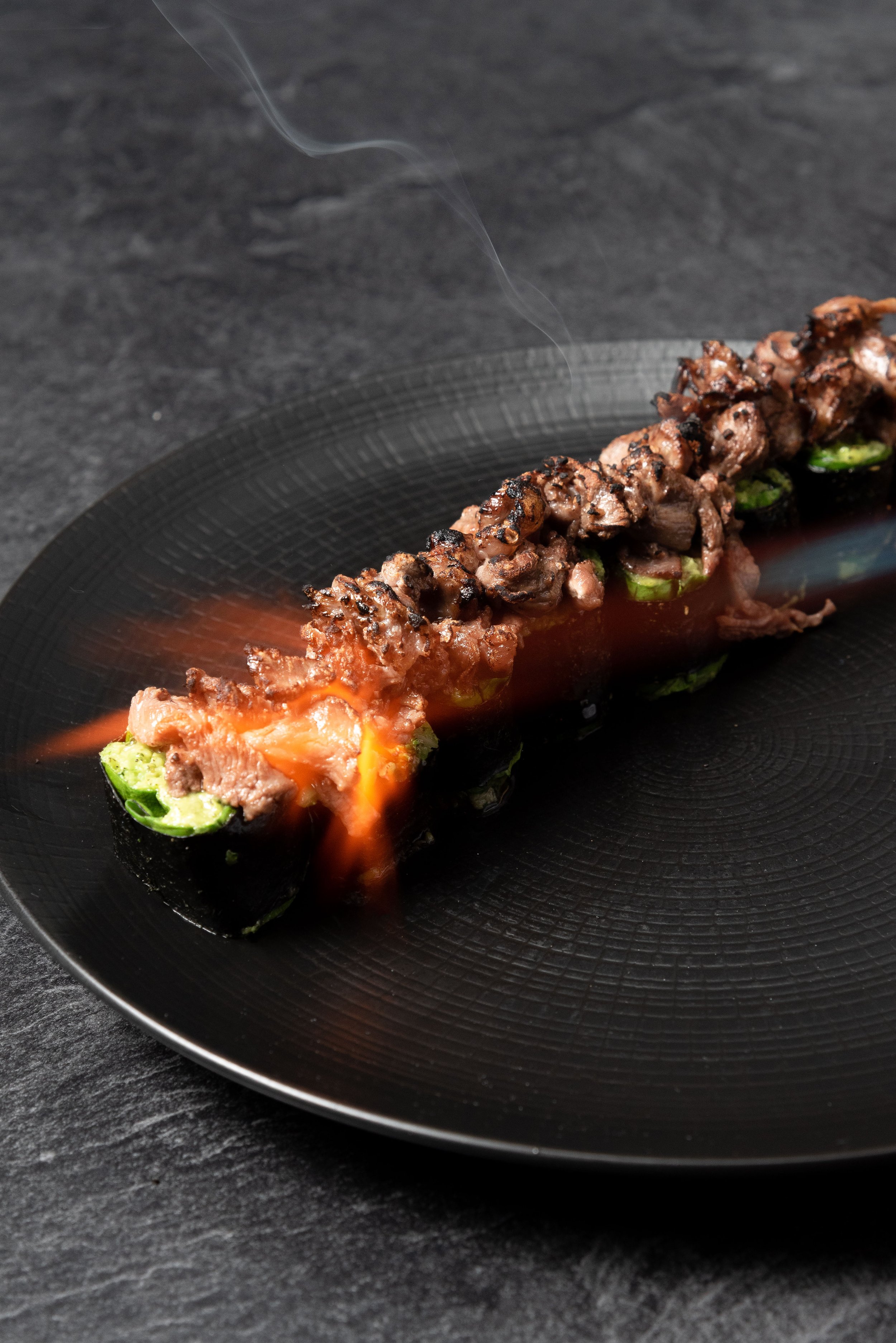 A black plate with a skewer of grilled meat and vegetables, with some flames and smoke rising from it.