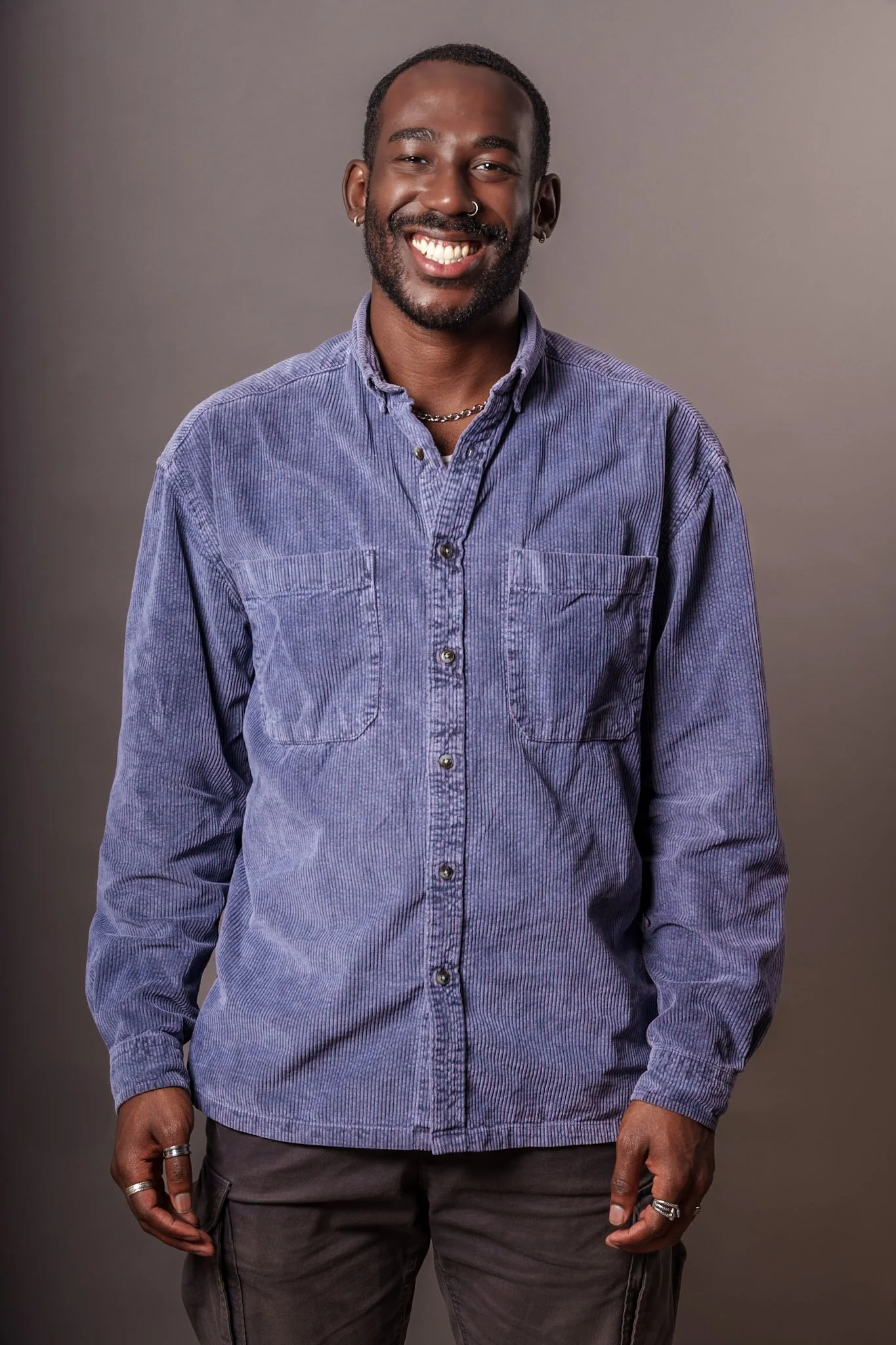 Portrait of a smiling Black man with short hair, wearing a blue corduroy button-up shirt, earrings, rings, and a chain necklace, standing against a neutral background.