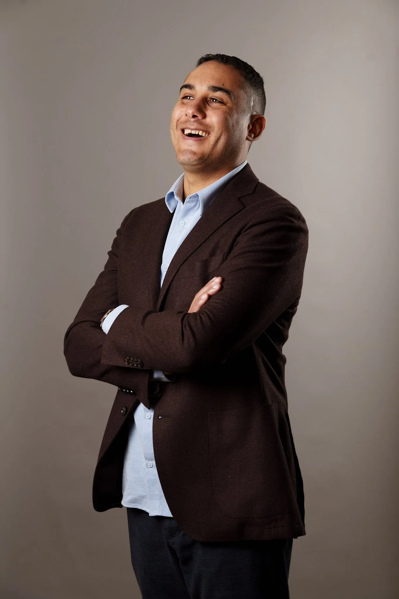 A confident man in a brown blazer and light blue shirt standing with arms crossed, smiling against a plain gray background.