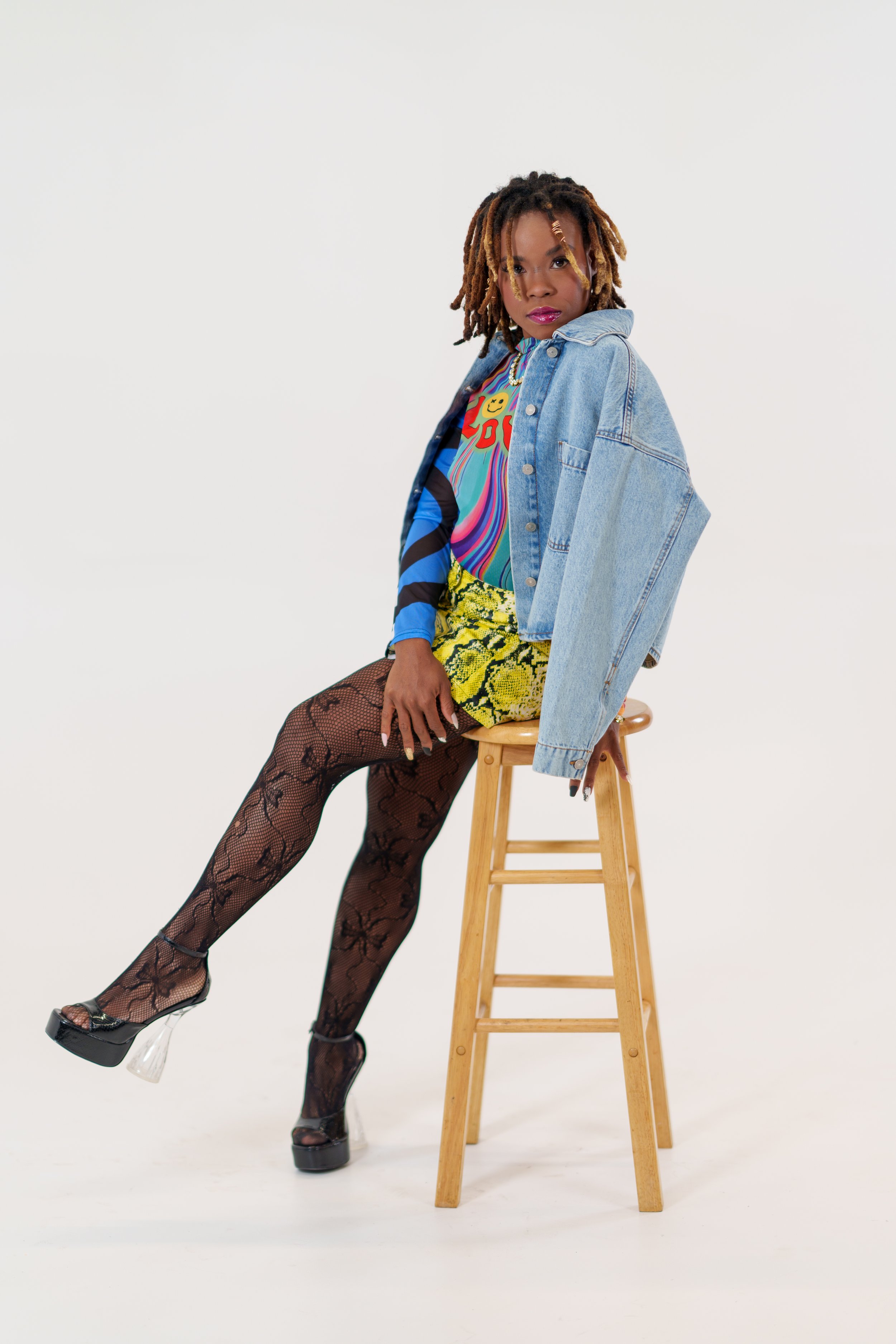 A woman with dreadlocks sitting on a wooden stool against a plain white background, wearing a colorful shirt, a denim jacket draped over her shoulder, a yellow snakeskin skirt, fishnet stockings, and high heels.