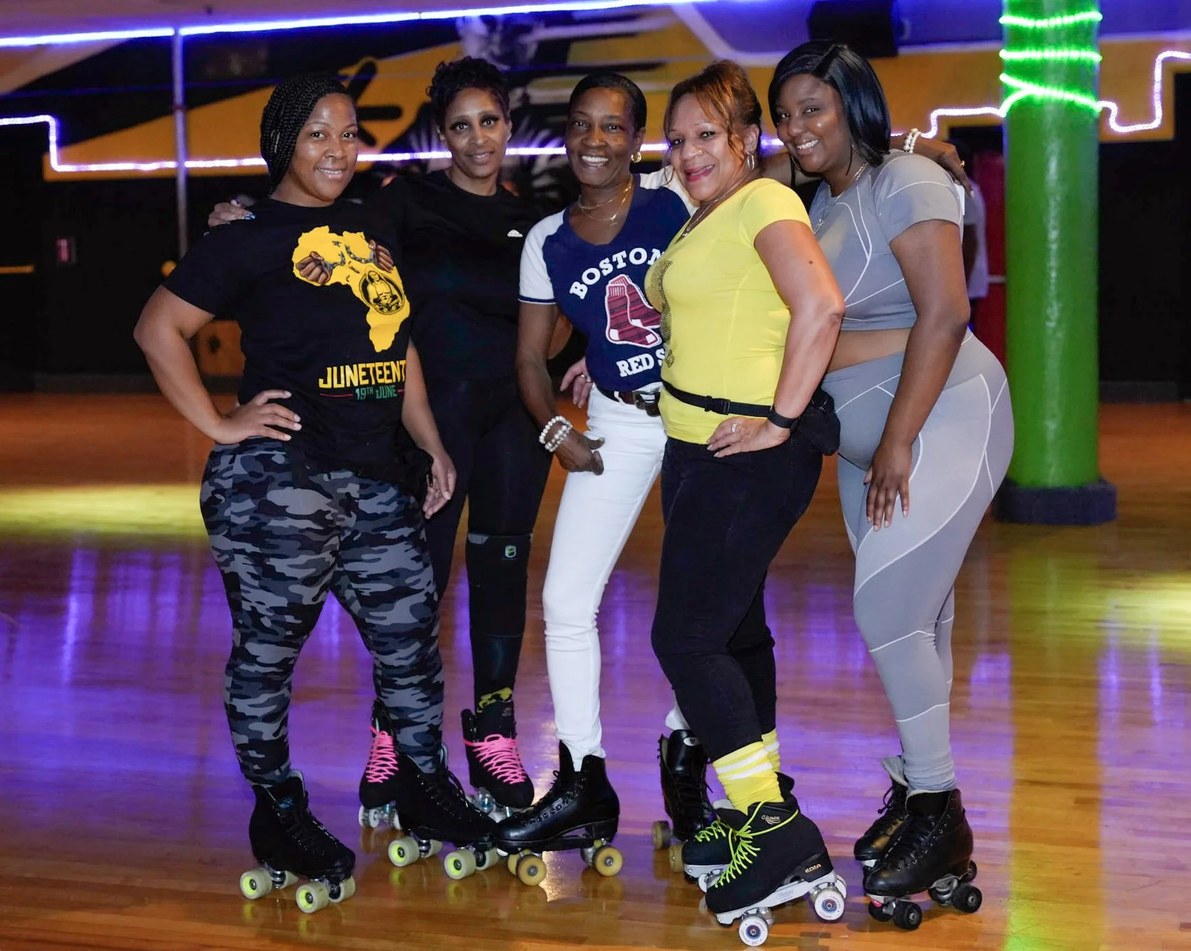 Five women on roller skates posing together in a roller rink with colorful neon lights.