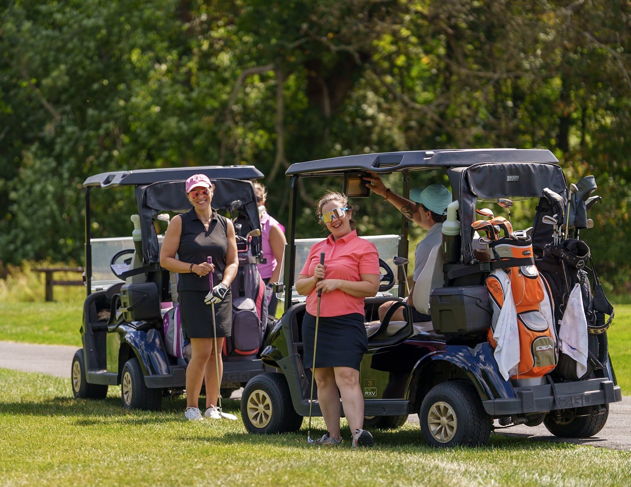 Four women standing and sitting around golf carts with golf bags on a grassy area at a golf course, with trees in the background.