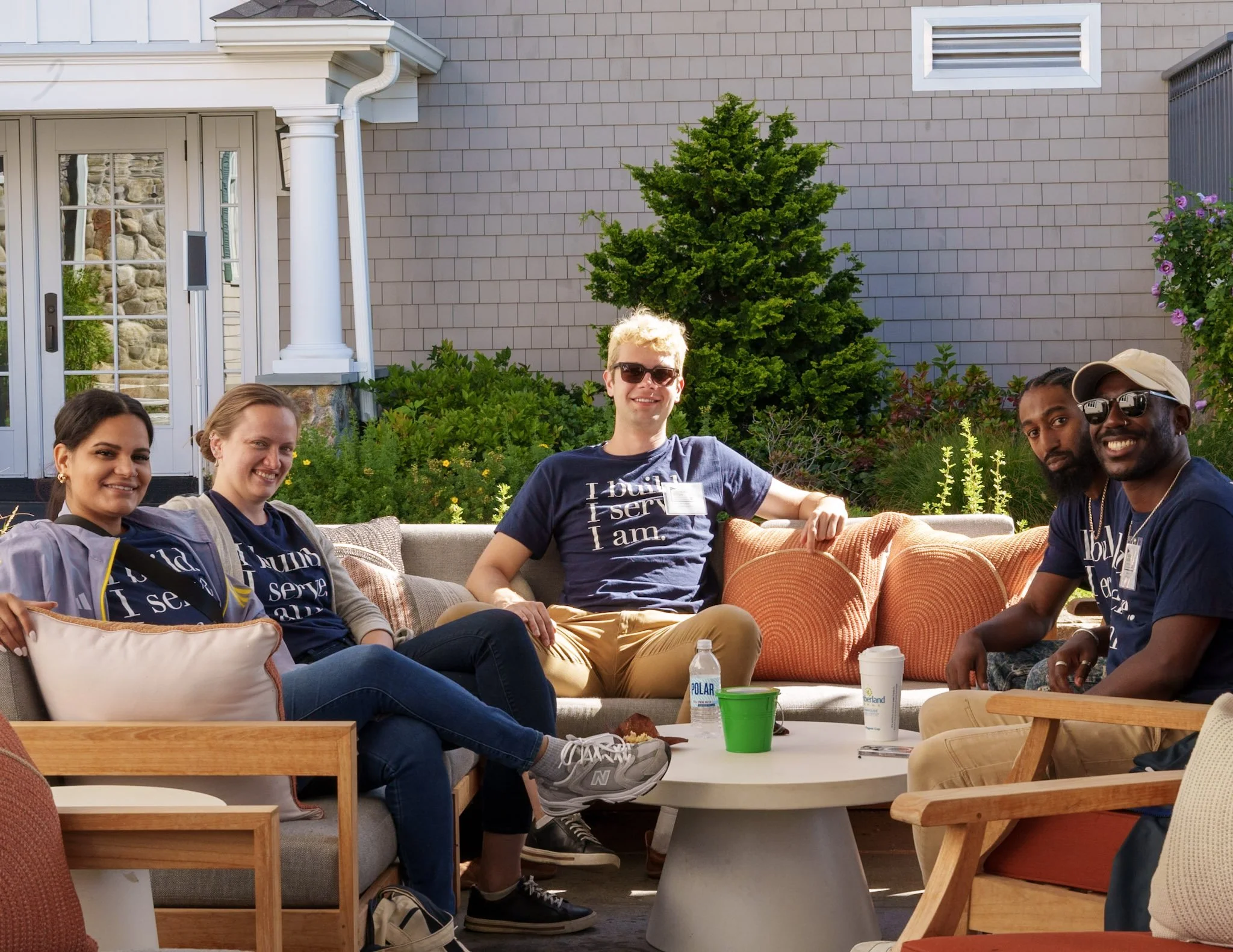 Group of five people sitting outdoors on a patio, smiling, with a house and garden in the background.