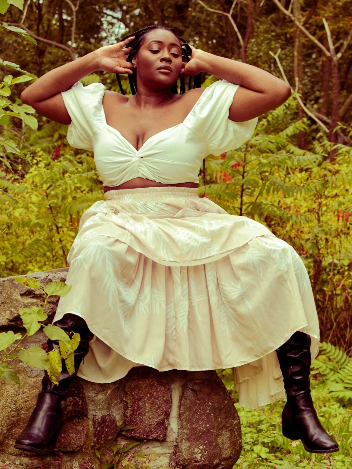 A woman with braided hair sitting on a stone, surrounded by green foliage, wearing a cream-colored blouse and flowy skirt with black boots.