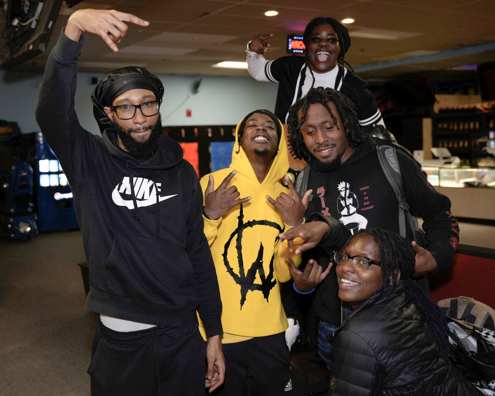 Group of six diverse young adults at an indoor arcade, posing and making expressive hand gestures, with bright smiles and playful gestures.