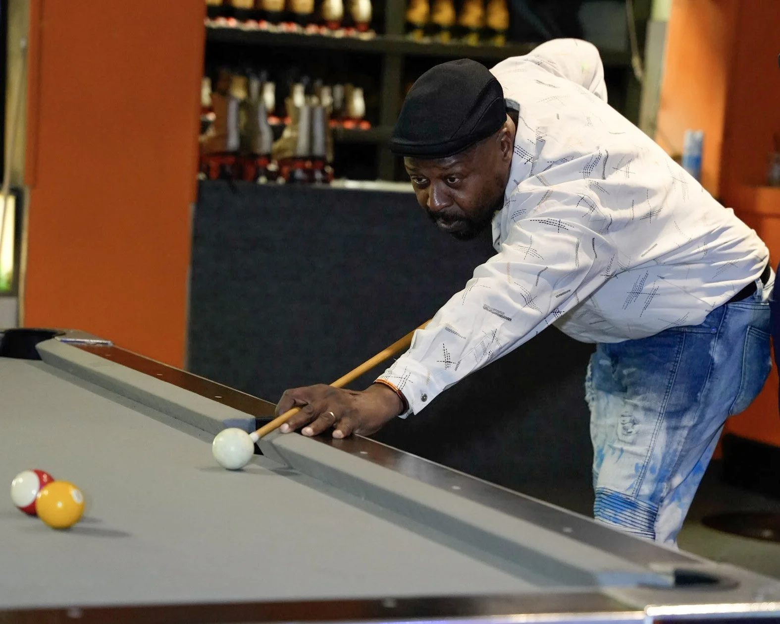 A man playing pool, aiming to hit the cue ball on a billiards table, wearing a white shirt, black cap, and ripped jeans in an indoor setting.