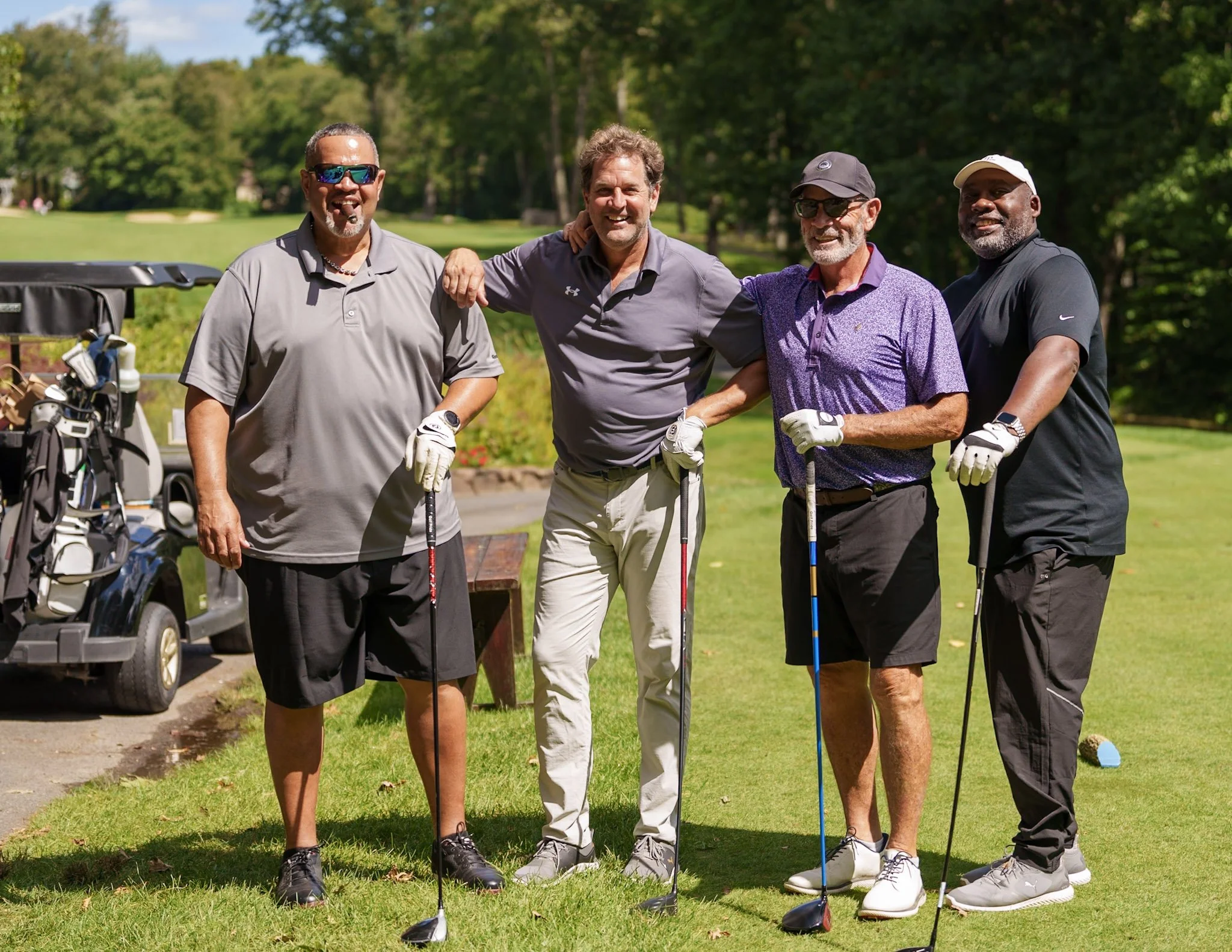 Four men standing on a golf course holding golf clubs, smiling, with a golf cart and trees in the background.