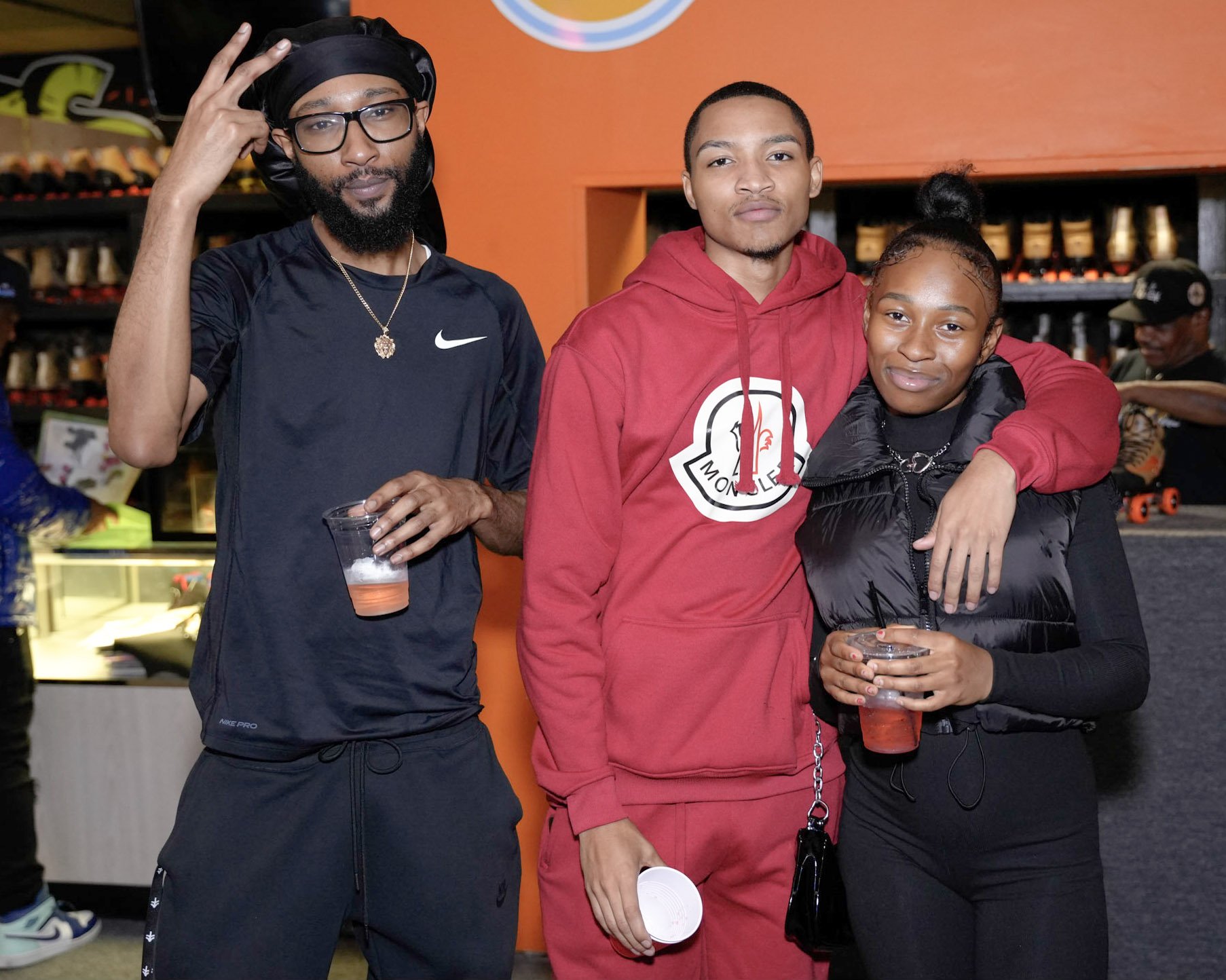 Three young adults standing together, smiling, with their arms around each other in a skateboarding shop or arcade. The person on the left wears glasses, a black Nike shirt, and a black headwrap, holding a cup of beverage. The person in the middle we