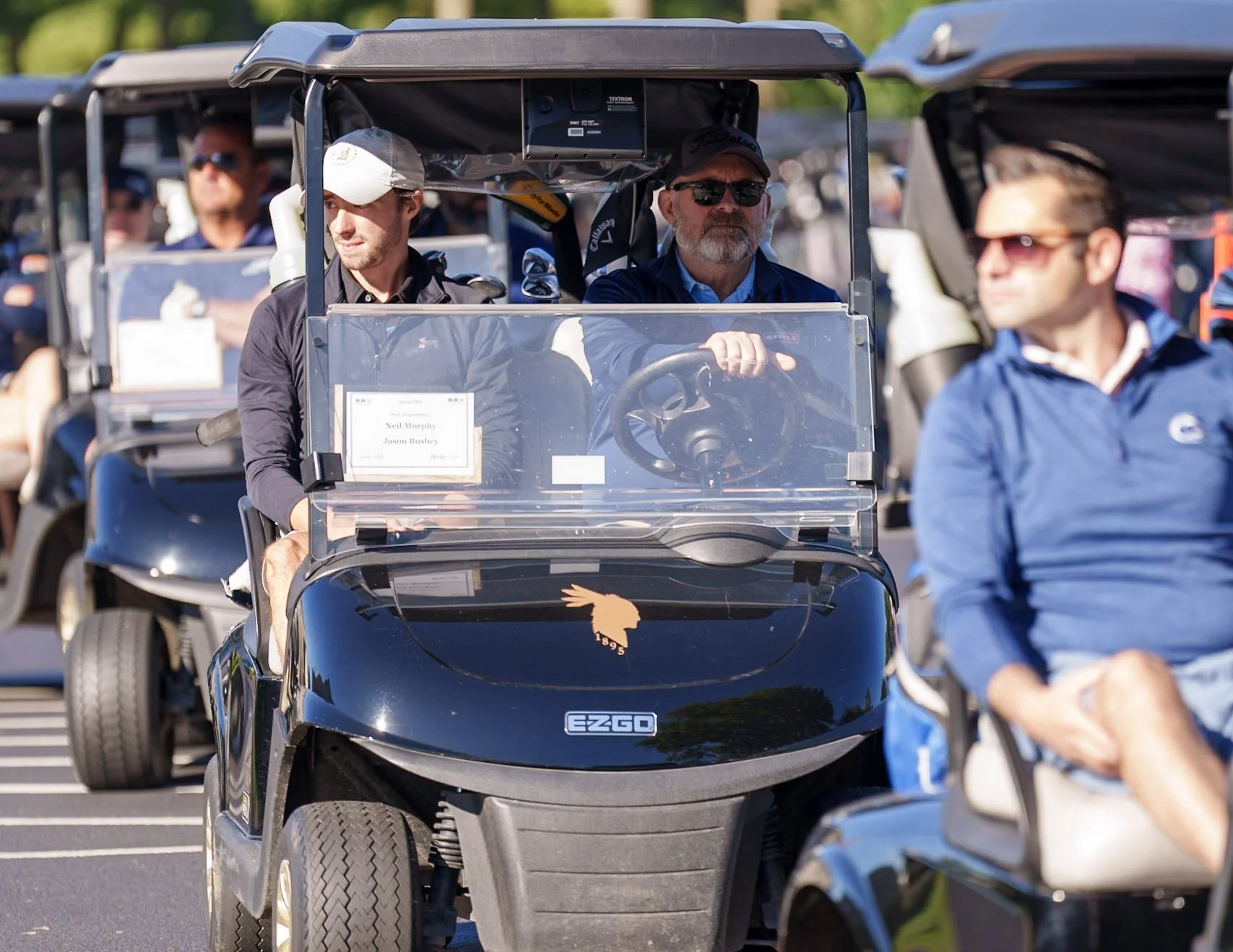 Golf carts lined up in a parking lot with people sitting inside, including a man in a black jacket and white cap in the front cart, and a man in a blue jacket with glasses in a separate cart, under bright sunlight with trees in the background.