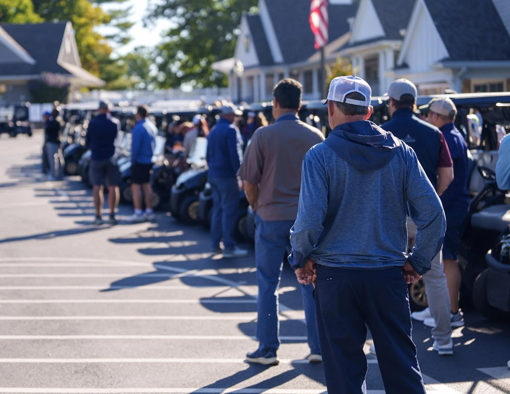 People standing in line outside at a golf course, with golf carts parked behind them and residential houses in the background.