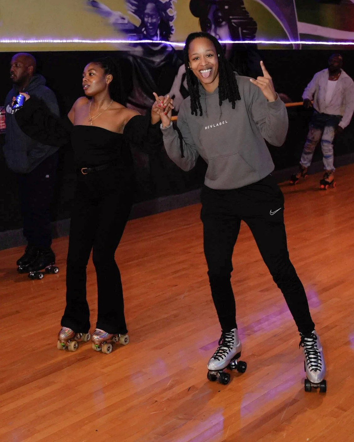 Two women roller skating on a wooden floor, holding hands and smiling at the camera, with other skaters in the background at an indoor skating rink.