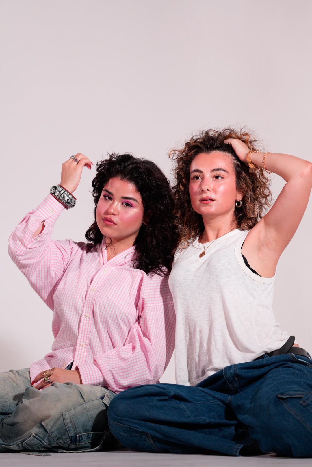 Two women sitting on the floor against a plain background, posing with relaxed expressions, one wearing a pink checkered shirt and the other in a white sleeveless top.