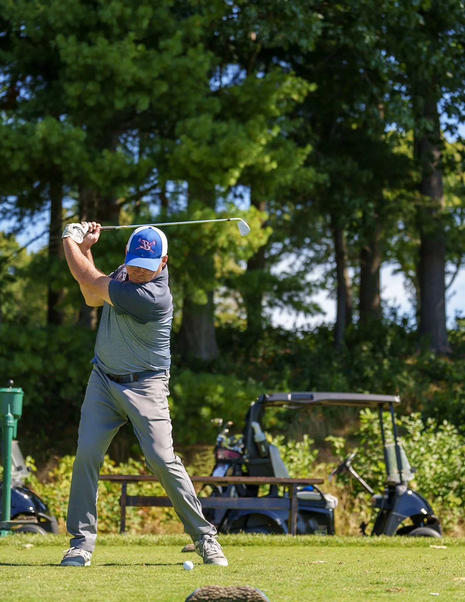 A man wearing a gray polo shirt, beige pants, a blue cap, and sunglasses is playing golf. He is swinging a golf club on a grassy golf course with green trees in the background. Two golf carts are parked behind him.