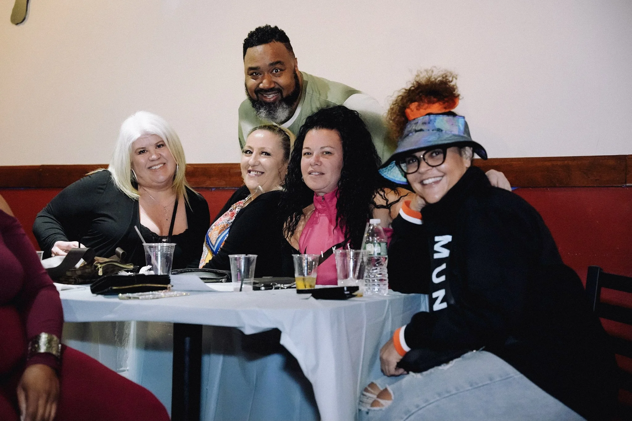 Group of five women and one man smiling at a social gathering, sitting at a table with drinks and food, one woman wearing a colorful hat, others dressed casually.