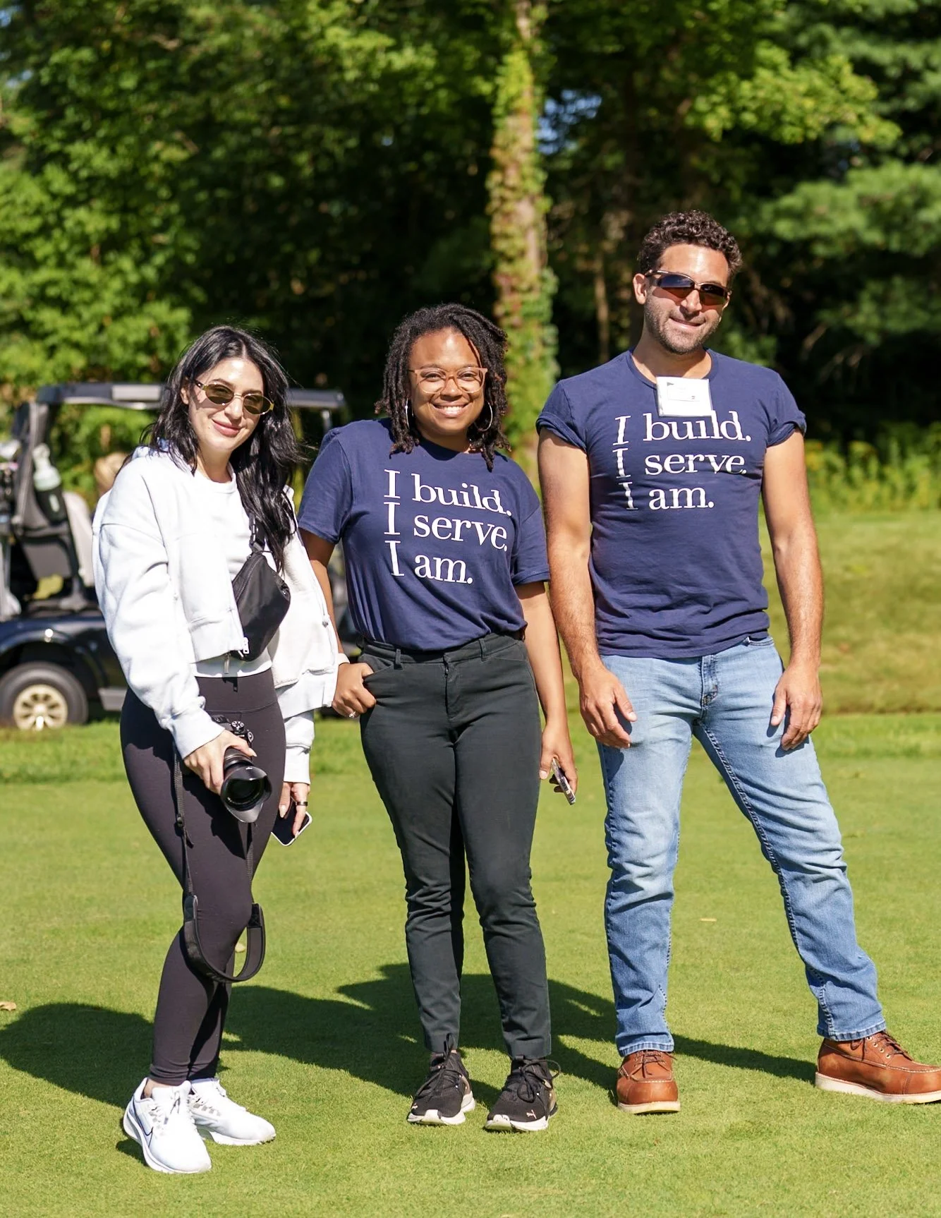 Three people standing on a golf course, smiling at the camera. Two women and one man, with the women wearing matching navy blue T-shirts that say 'I build. I serve. I am.'.
