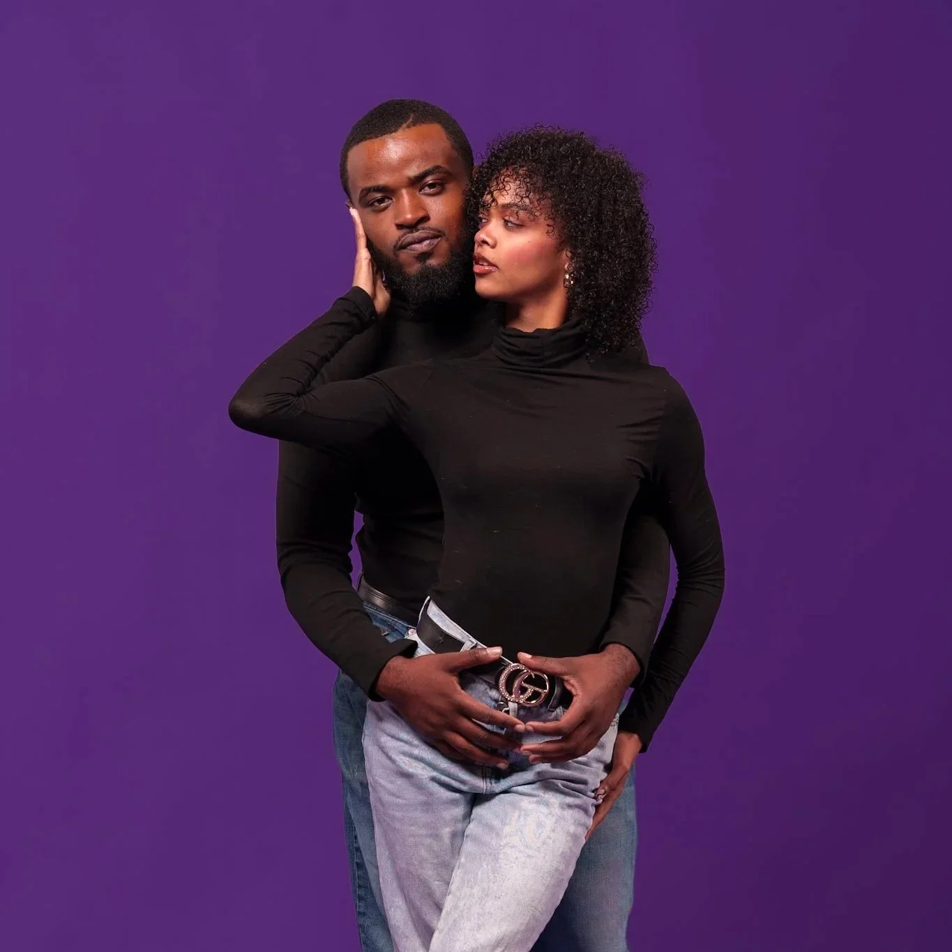 A man and woman pose together against purple background, with the man behind and the woman in front, both wearing black tops.
