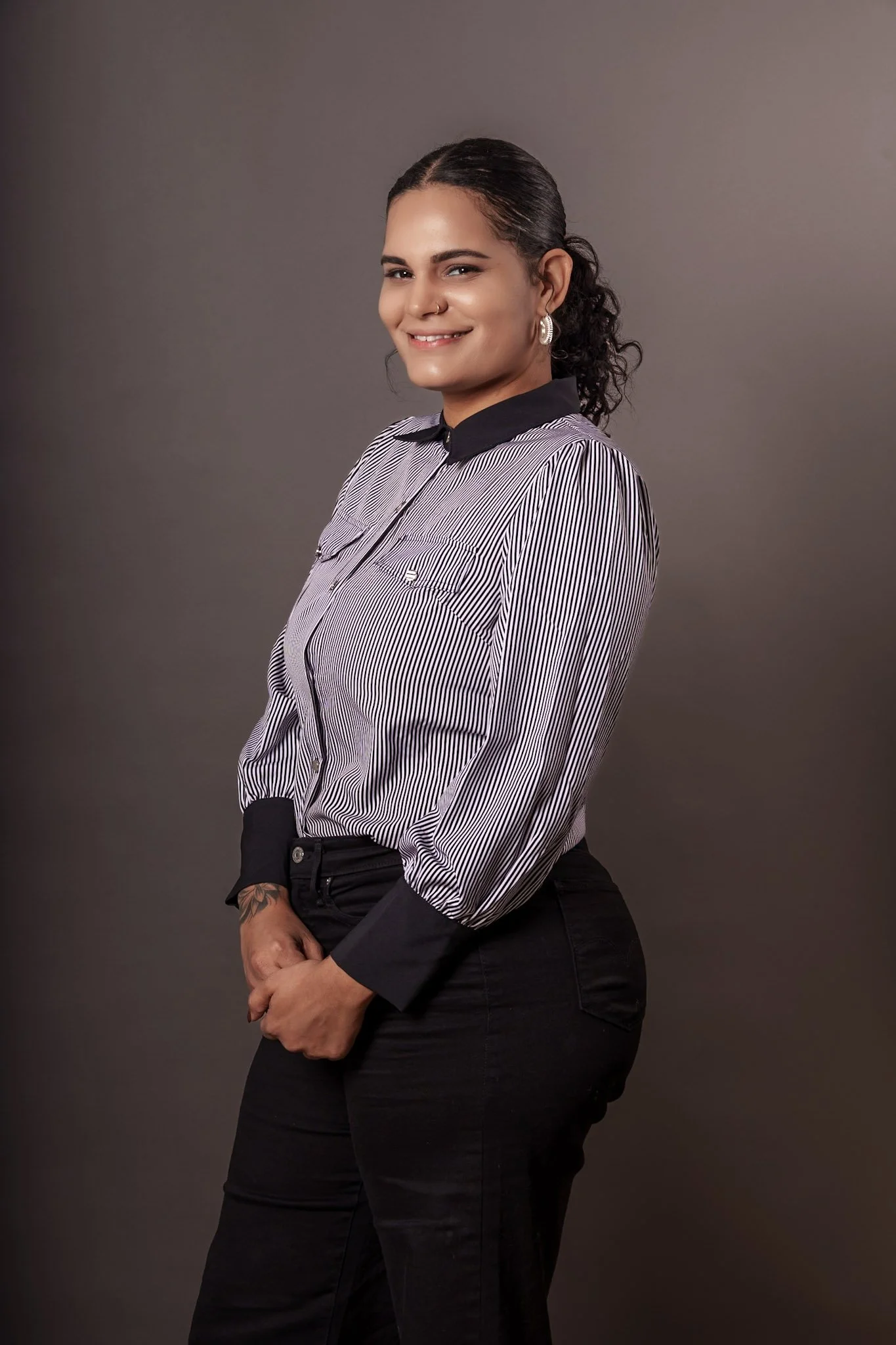 A young woman with curly dark hair pulled back, wearing a striped blouse with black collar and cuffs, black pants, standing against a plain background, smiling and looking at the camera.