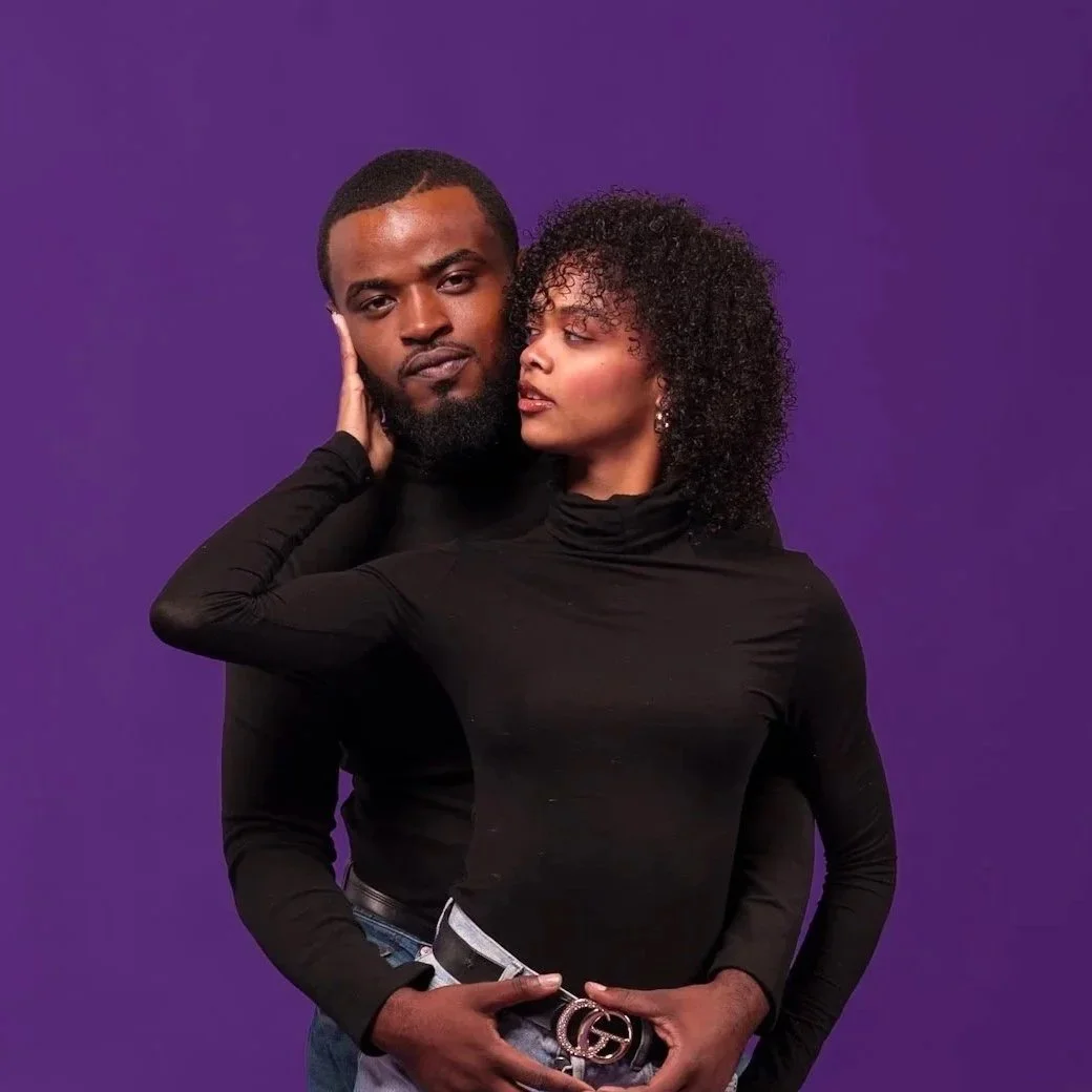 A man and a woman posed closely together against a purple background. The woman is touching the man's face gently, and they are both looking at each other. The man has a beard and short hair, and the woman has curly hair and wears a black turtleneck.