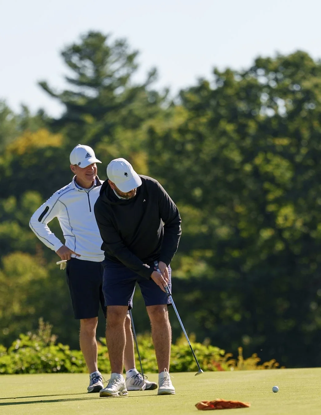 Two men playing golf on a sunny day, with one putting and the other watching, on a green with trees in the background.