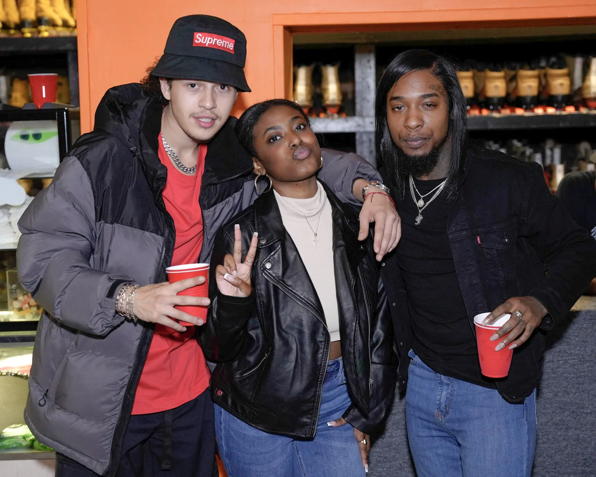 Three young adults posing together at a party or gathering, holding red cups, with a colorful background.