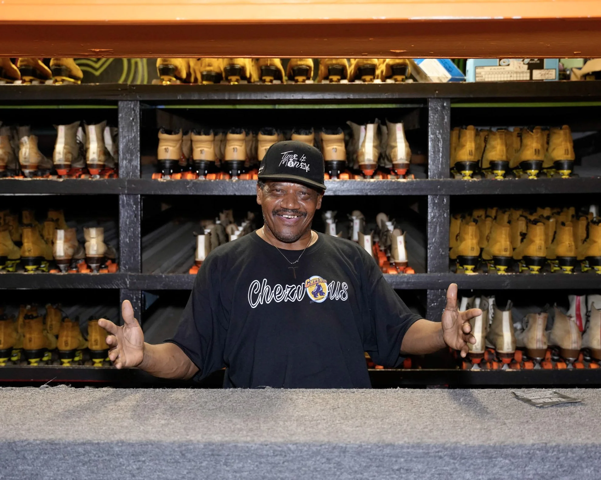 A man smiling and gesturing with his hands standing in front of a shelf filled with yellow roller skates.