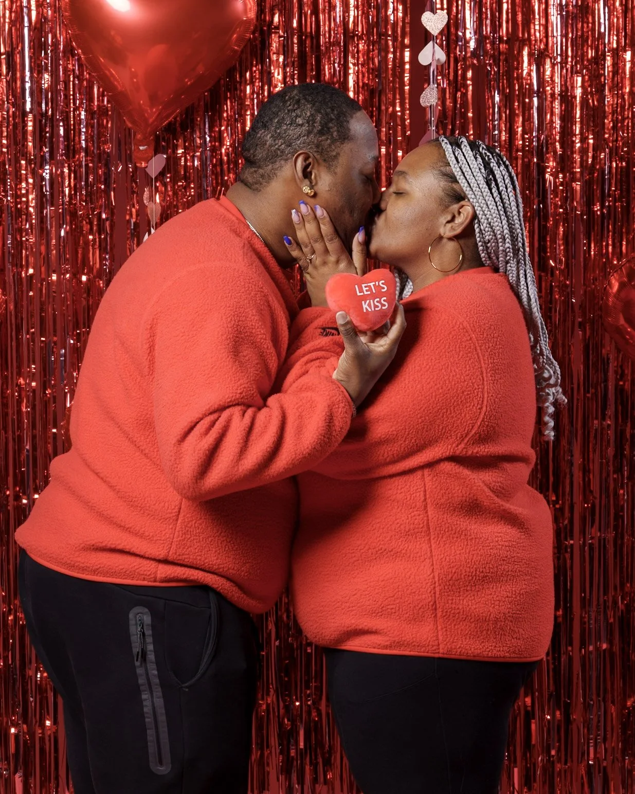 Couple dressed in red fleece jackets sharing a kiss in front of a red tinsel backdrop, with heart-shaped balloons and a small red heart pillow that says 'Let's Kiss'.