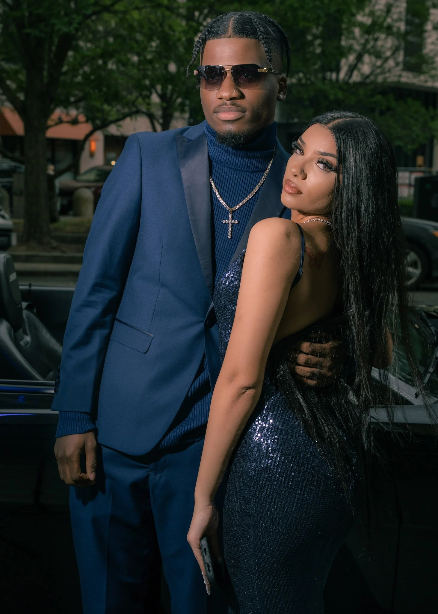 A man in a blue suit and sunglasses standing next to a woman in a sparkly black dress with long black hair, outdoors with trees and cars in the background.