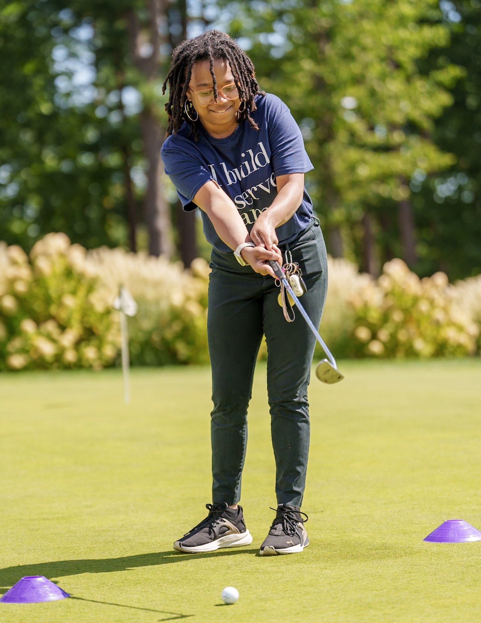 A woman with glasses and dreadlocks playing golf on a sunny day, wearing a navy blue shirt and black pants, focusing on her shot near a golf ball on the green, surrounded by trees and bushes.