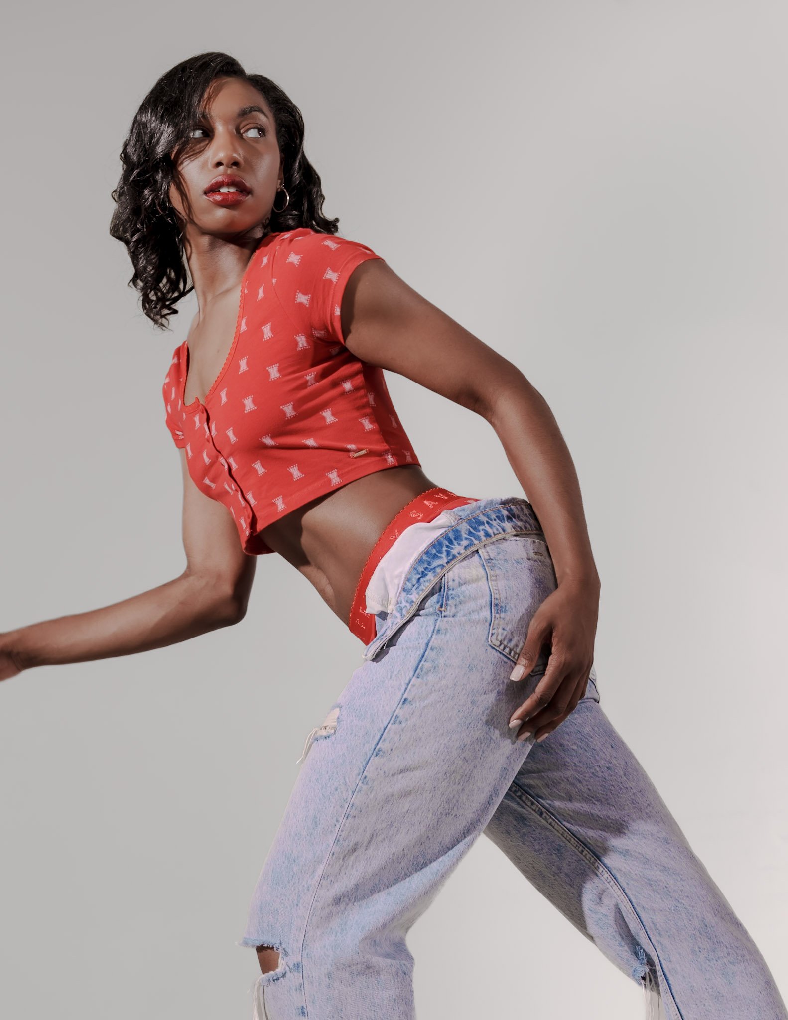 A young woman with dark, wavy hair poses against a plain background, wearing a red cropped top with a pattern, distressed light Denim jeans, and hoop earrings.