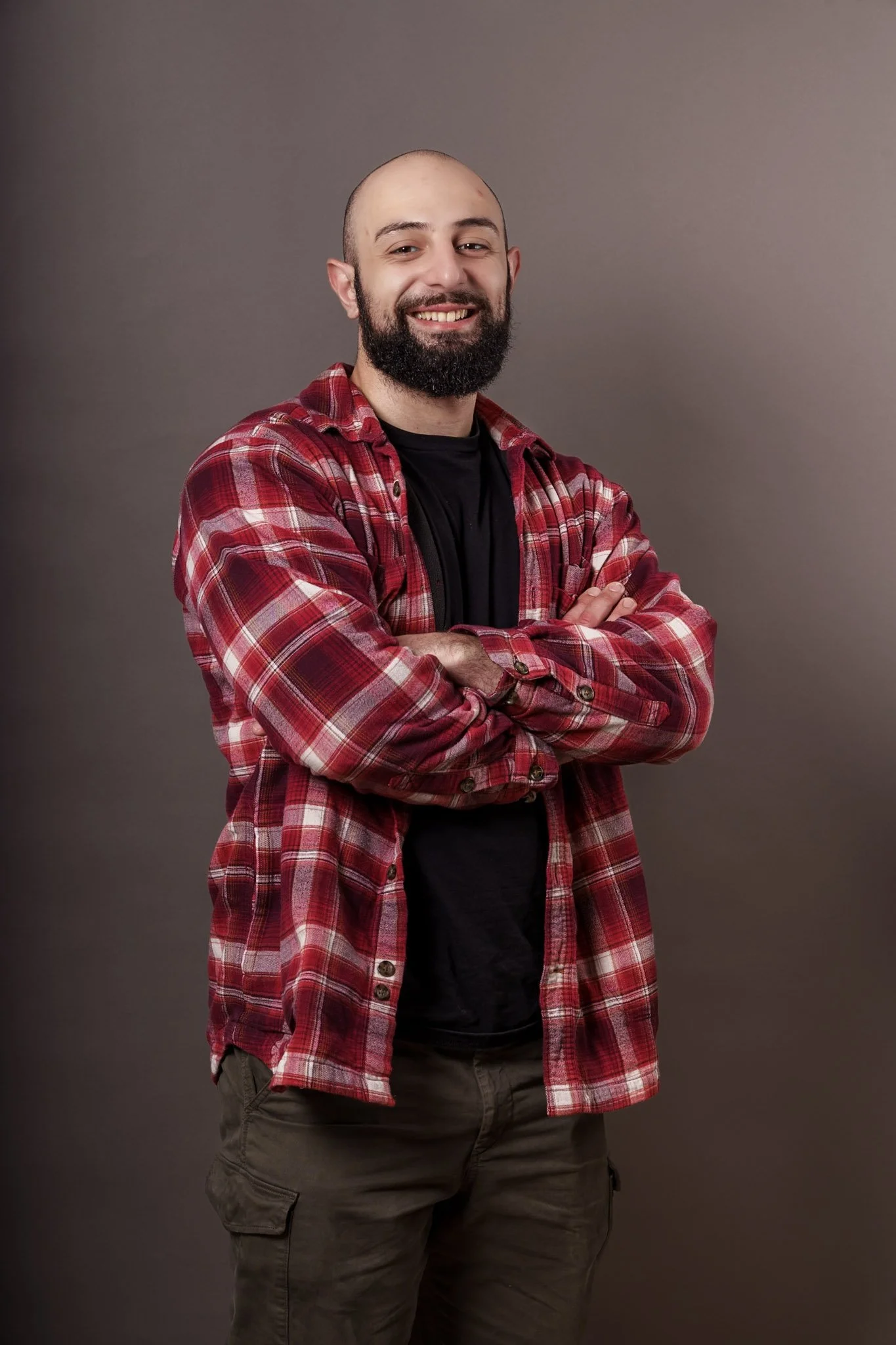 A man with a beard and bald head smiling with arms crossed, wearing a red plaid shirt over a black t-shirt, standing against a gray background.