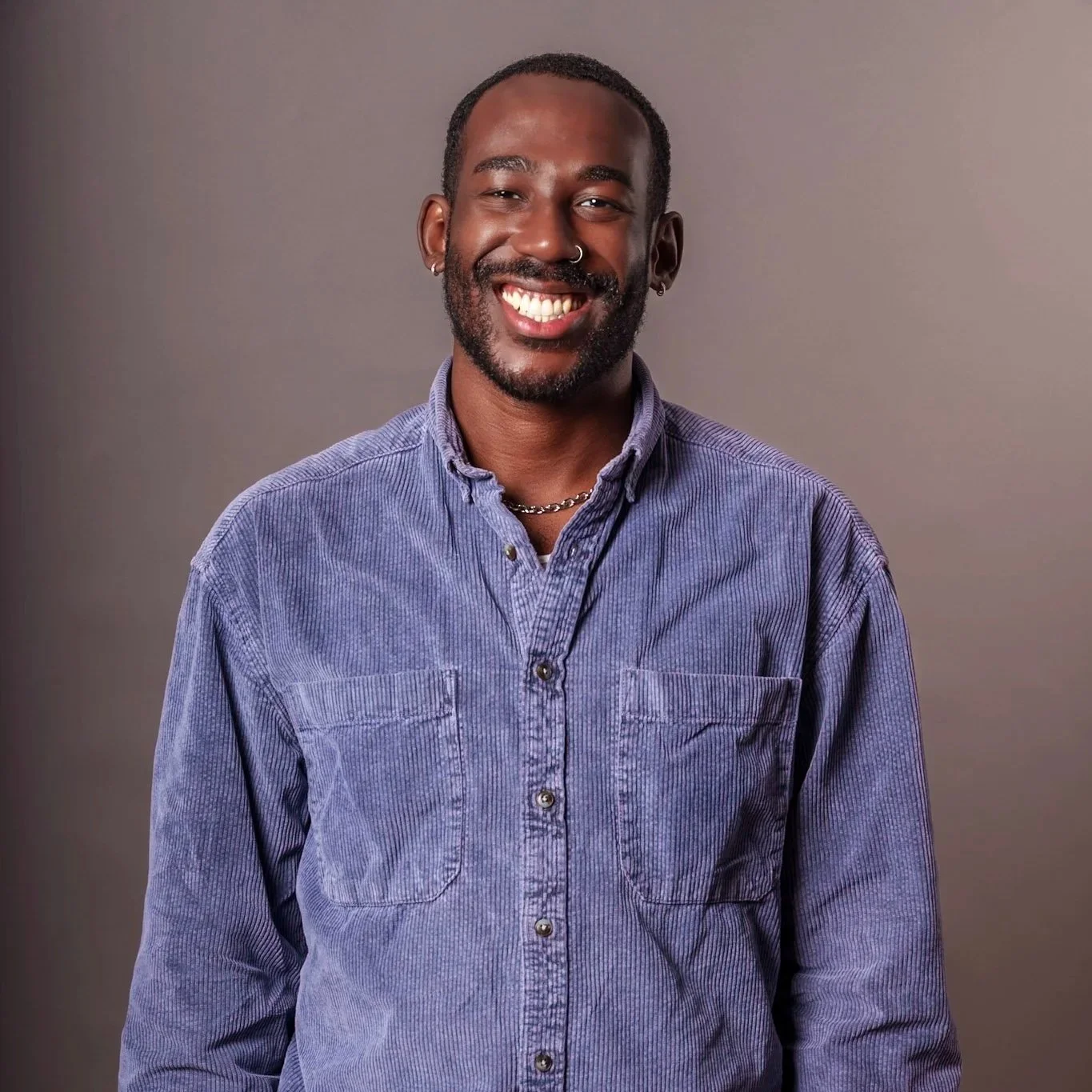 A smiling man with a beard and short hair, wearing a purple corduroy shirt and silver jewelry, standing against a neutral background.
