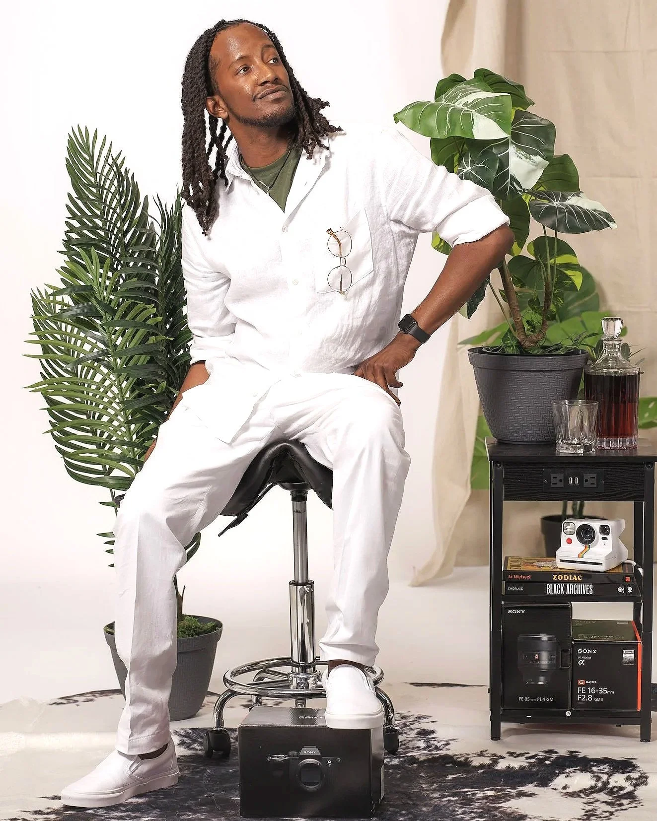Man with long dreadlocks and glasses hanging from his white shirt, wearing all white, sitting on a stool in front of a white background with green potted plants and black furniture with books and camera equipment.