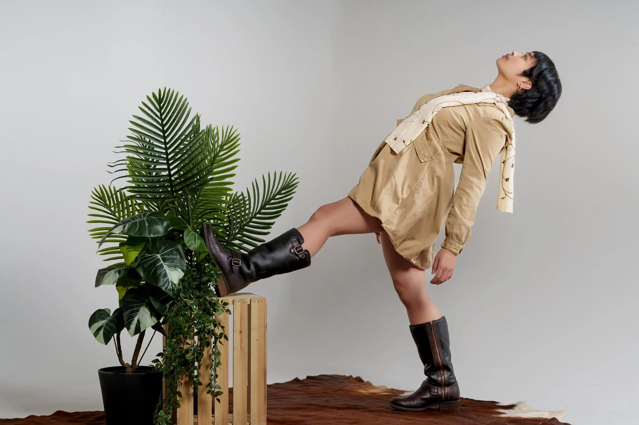 Person leaning back with one foot elevated on a wooden crate, next to a large green plant, wearing a tan jacket and knee-high leather boots, posing against a plain gray wall.