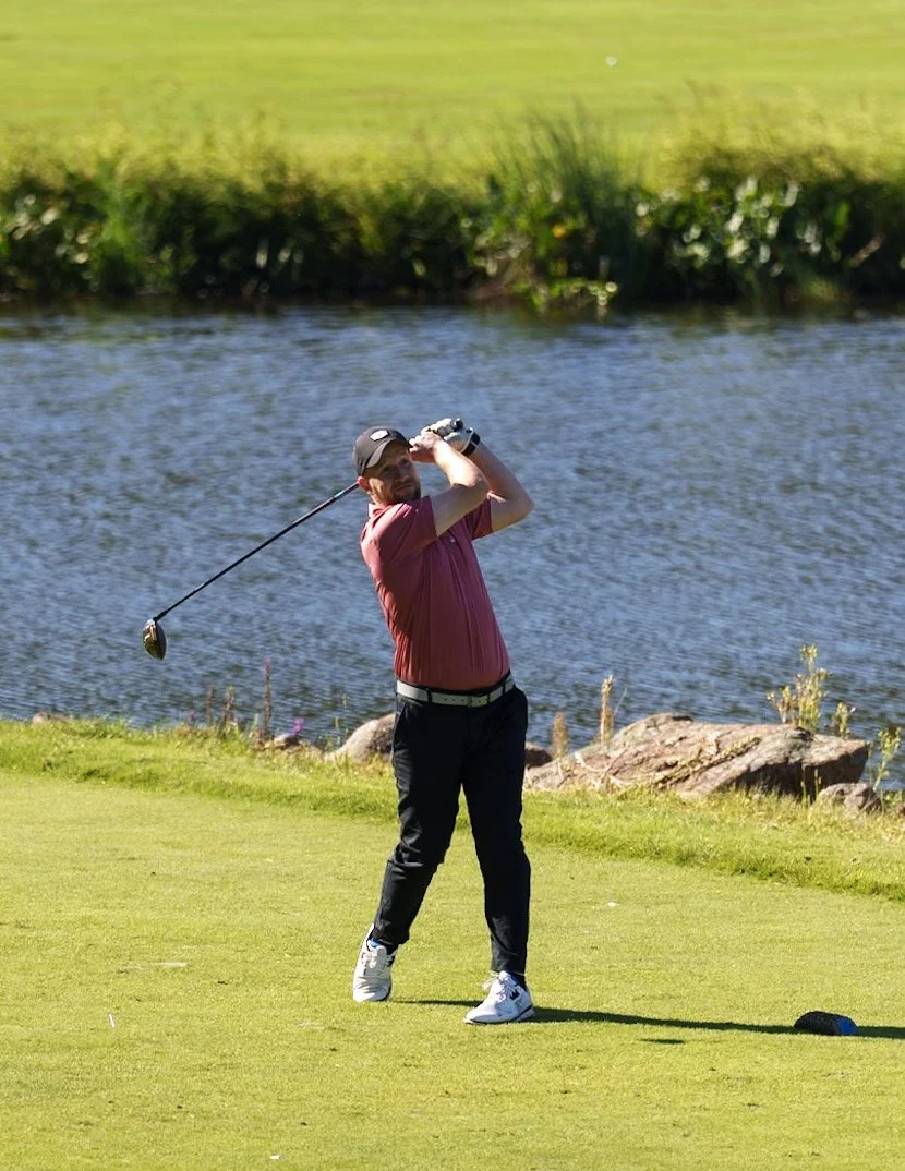 A man in a red shirt and black pants playing golf near a body of water on a sunny day.