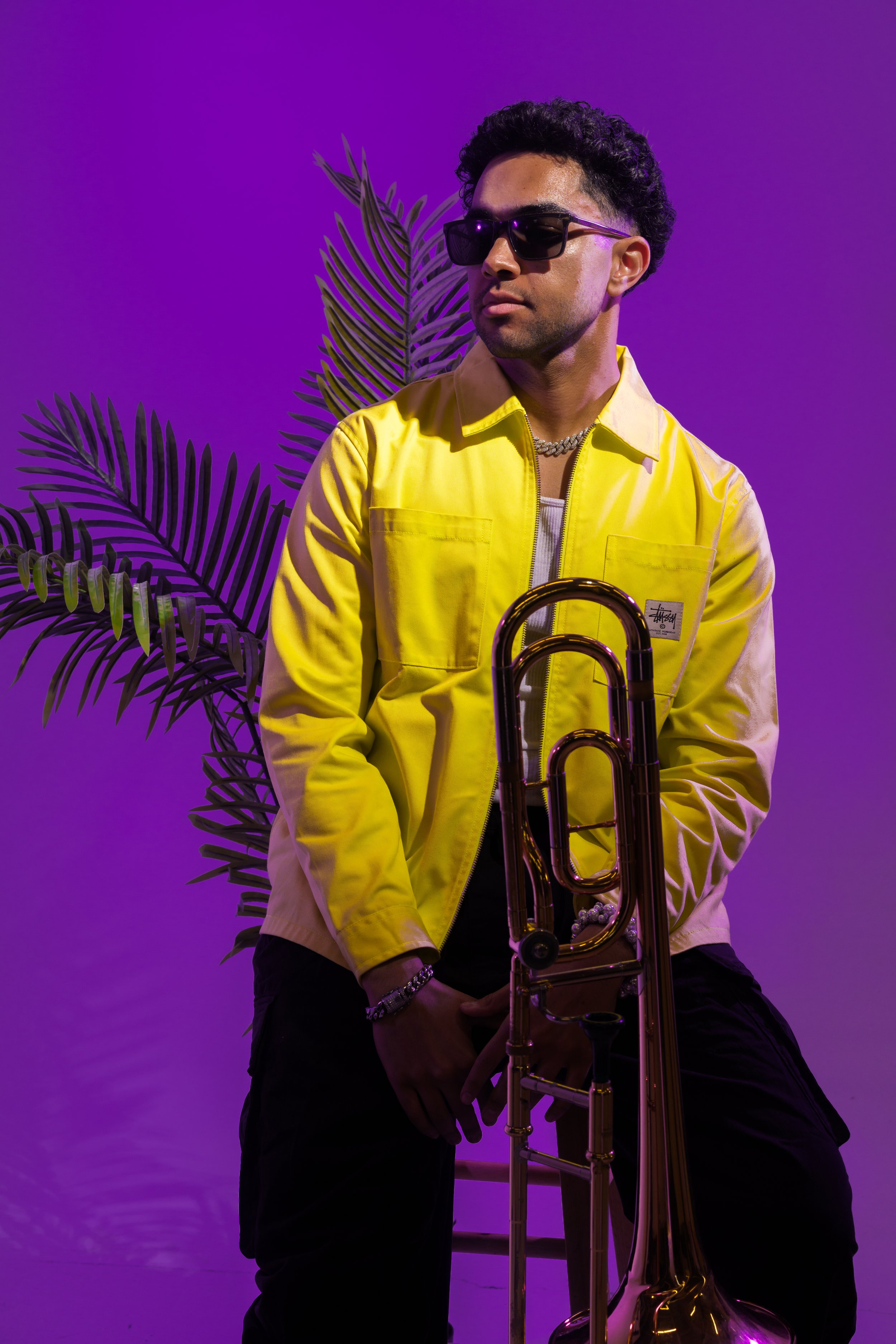 A man wearing sunglasses, a yellow jacket, and jewelry holding a trombone against a purple background with a plant.