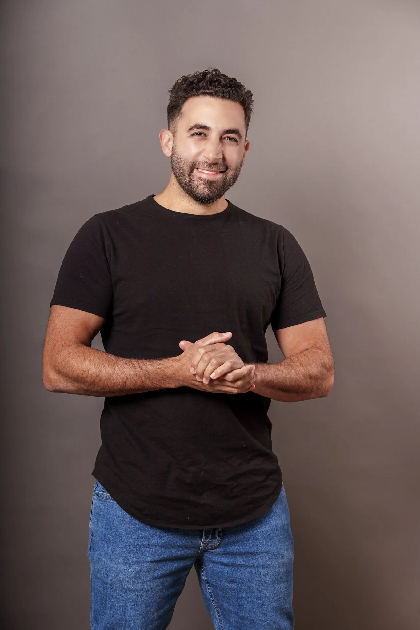 A man with dark hair and a beard, smiling, standing in front of a plain gray background, wearing a black t-shirt and blue jeans.