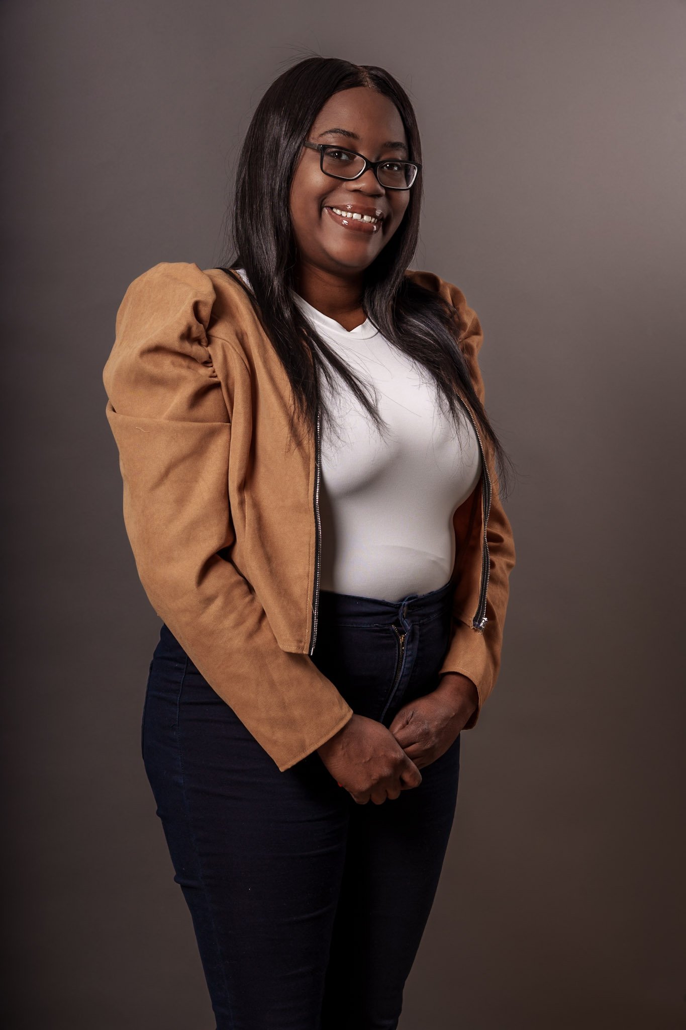 A woman with long black hair, glasses, and a warm smile, wearing a white top, brown jacket with puffed sleeves, and black pants, standing against a neutral background.