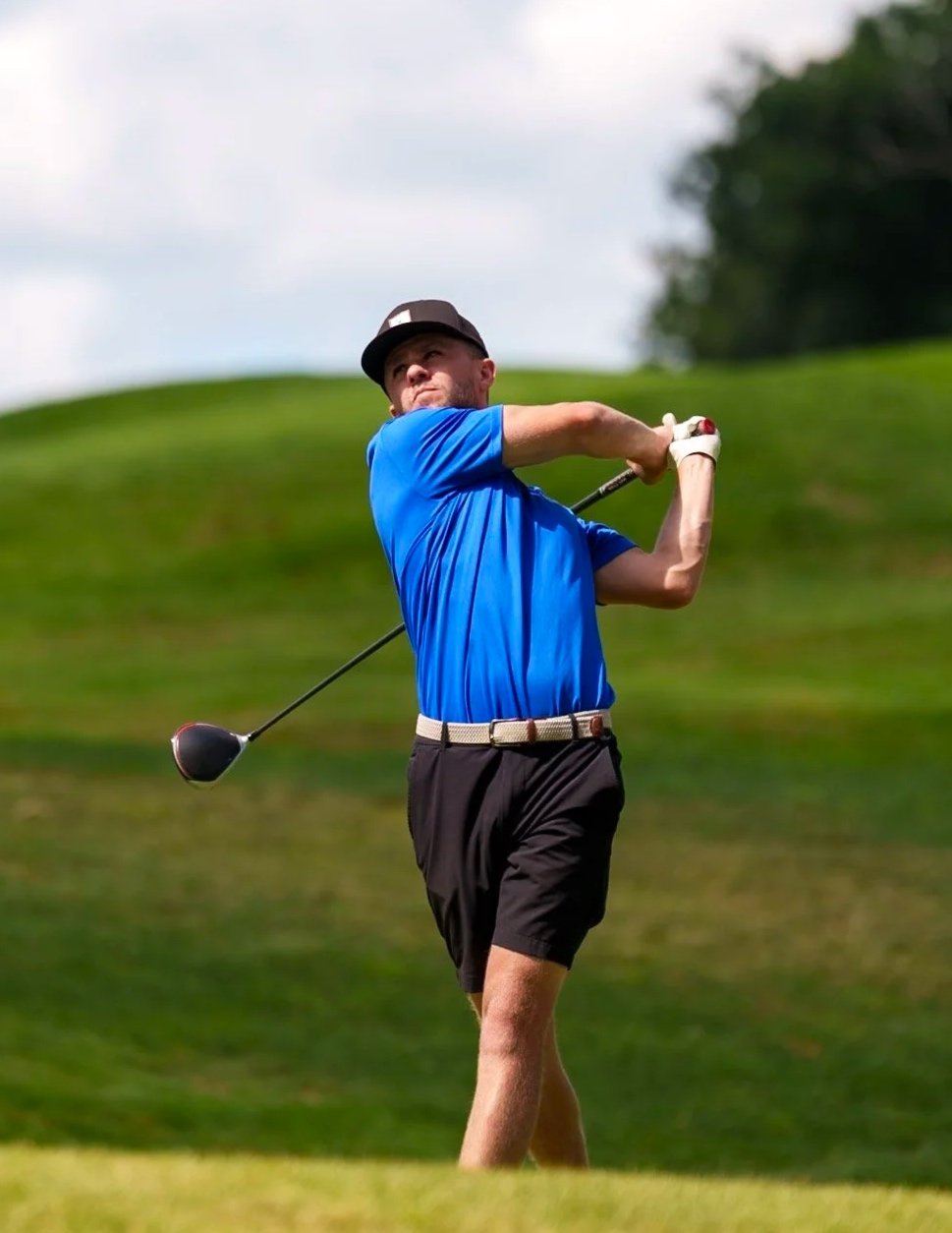 A man in a blue shirt and black shorts swinging a golf club on a golf course with green grass and trees in the background.
