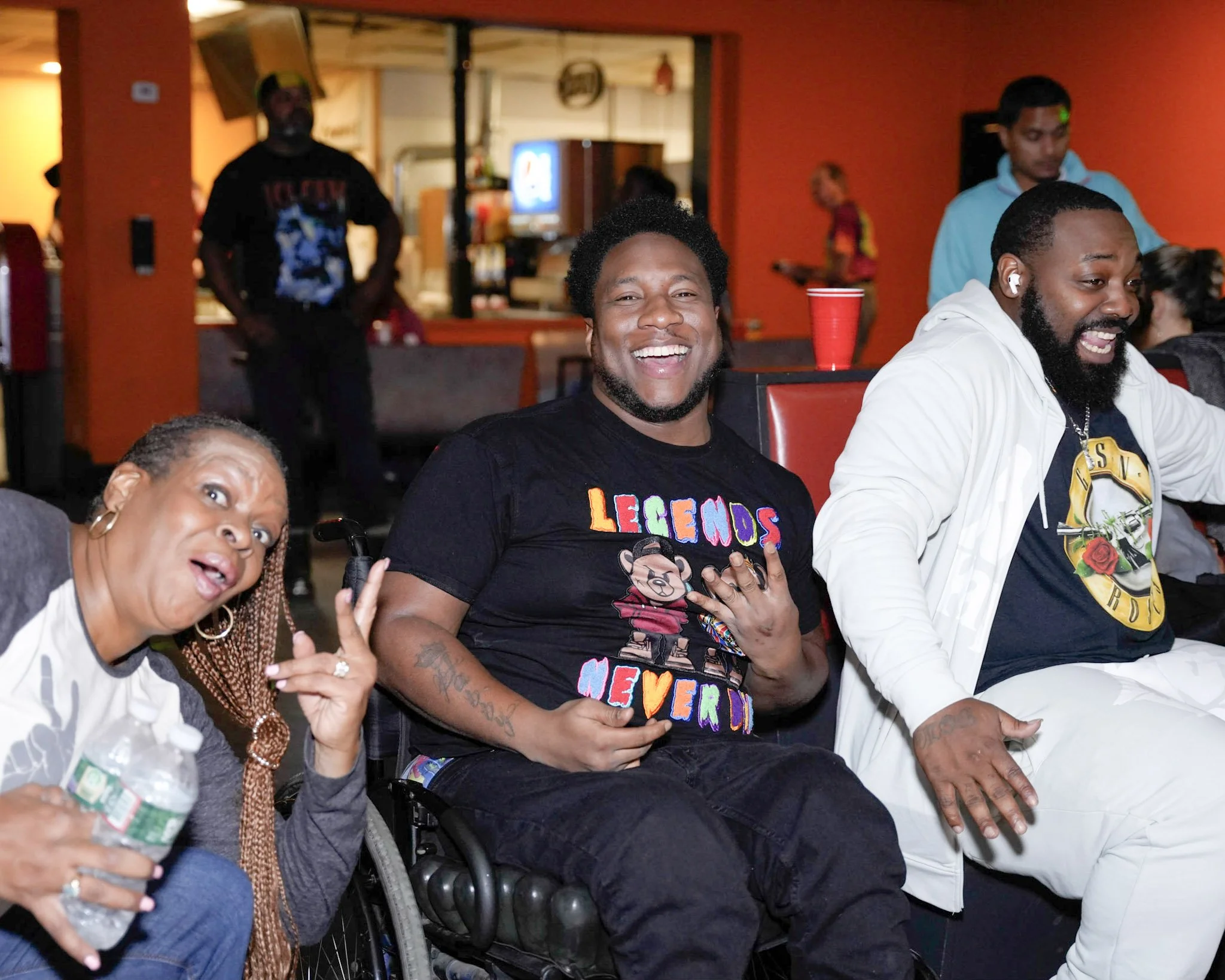 Three people sitting and smiling at a social gathering, one woman and two men, with one woman holding a water bottle, the man in the middle wearing a black shirt with colorful text, and the man on the right wearing a white jacket, in a lively room wi