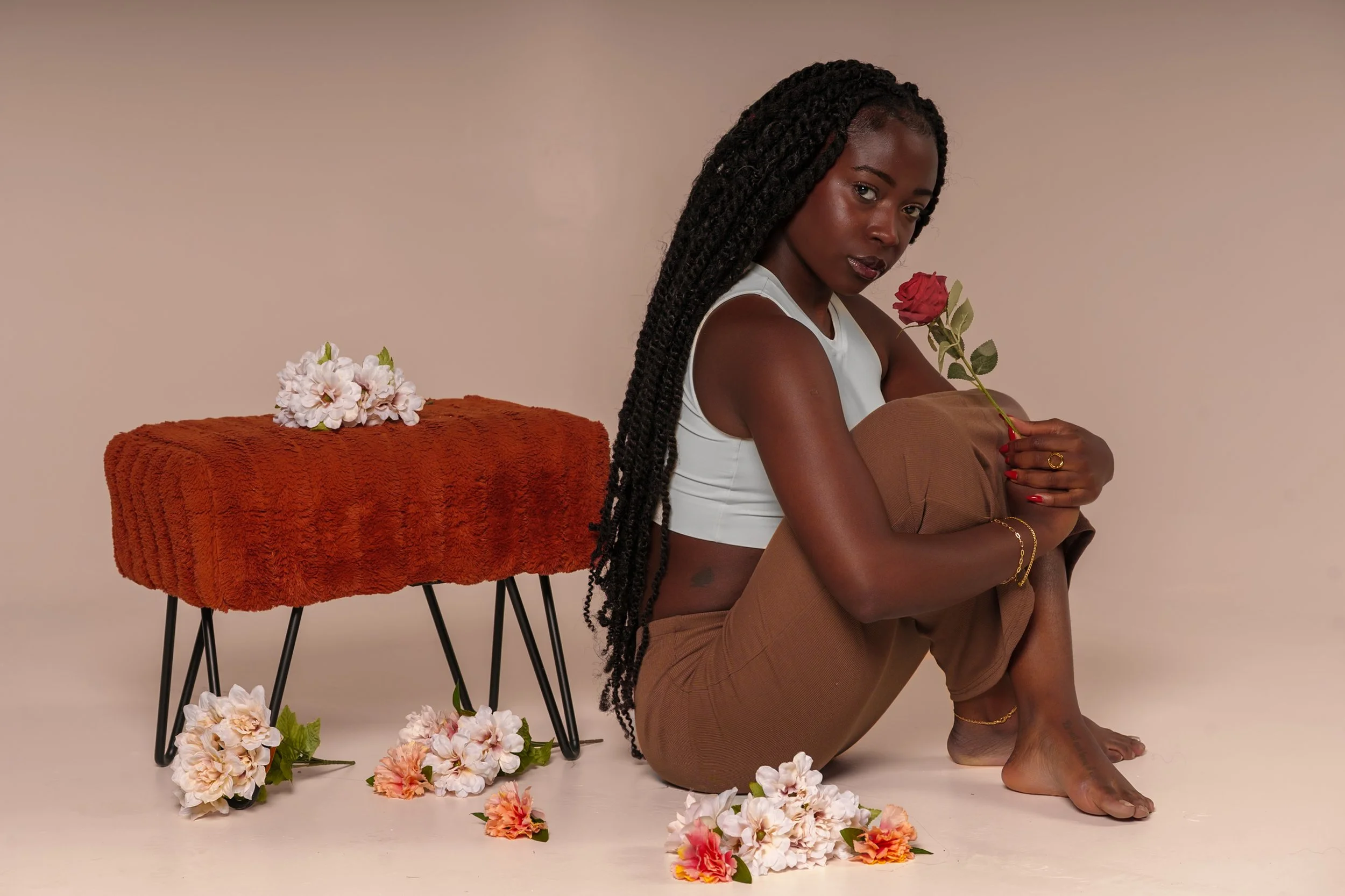 A woman with long braided hair, wearing a white crop top and brown pants, is sitting barefoot on the floor, holding a pink rose. She is surrounded by scattered flowers and is next to a rust-colored textured table with more flowers on and around it, s