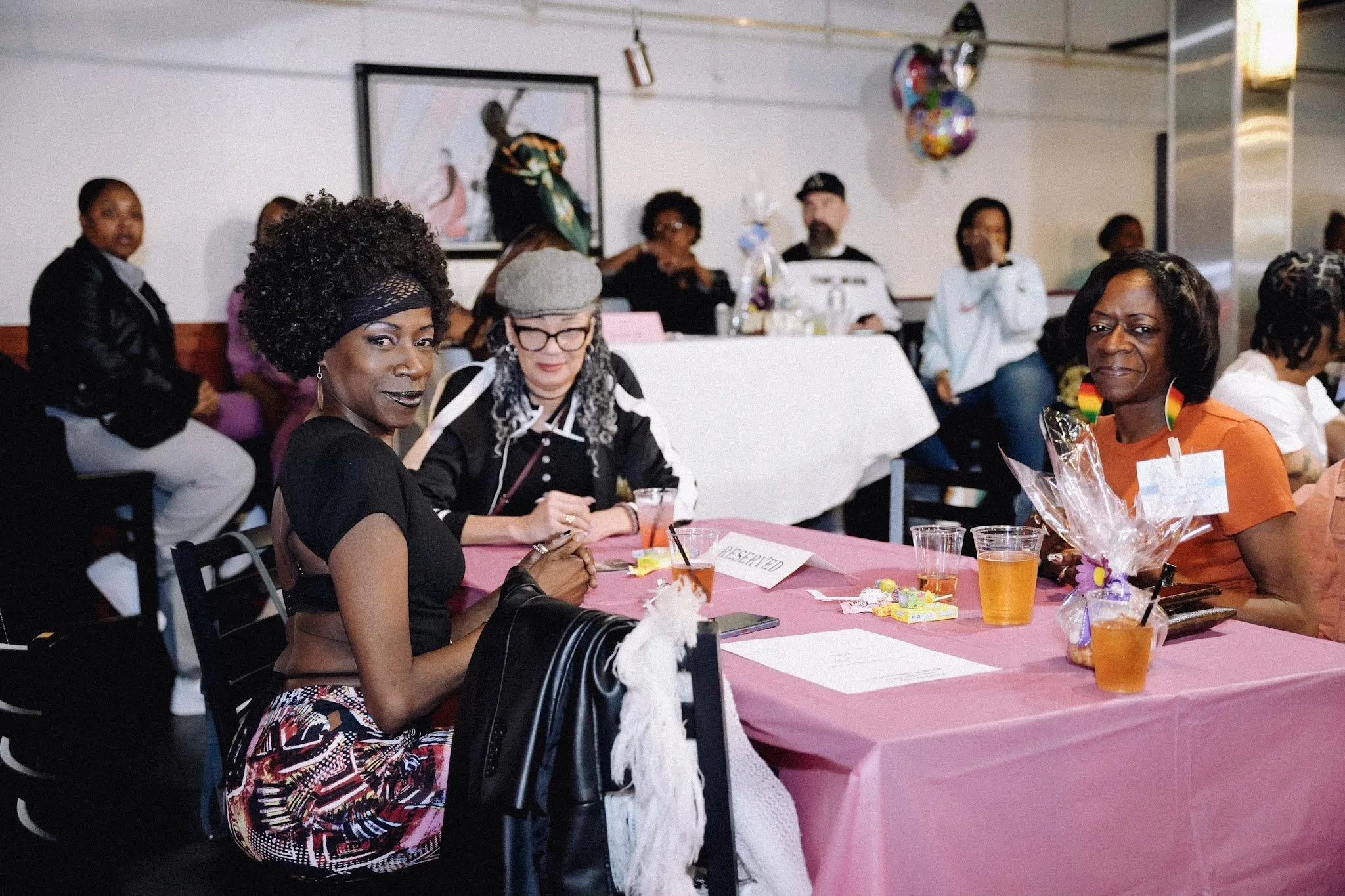 A group of women sitting at a long pink table during a social event or gathering. Some women are looking at the camera, while others are engaged in conversation or looking away. The table has drinks, gifts, and a name tag that reads 'RESERVED'. There