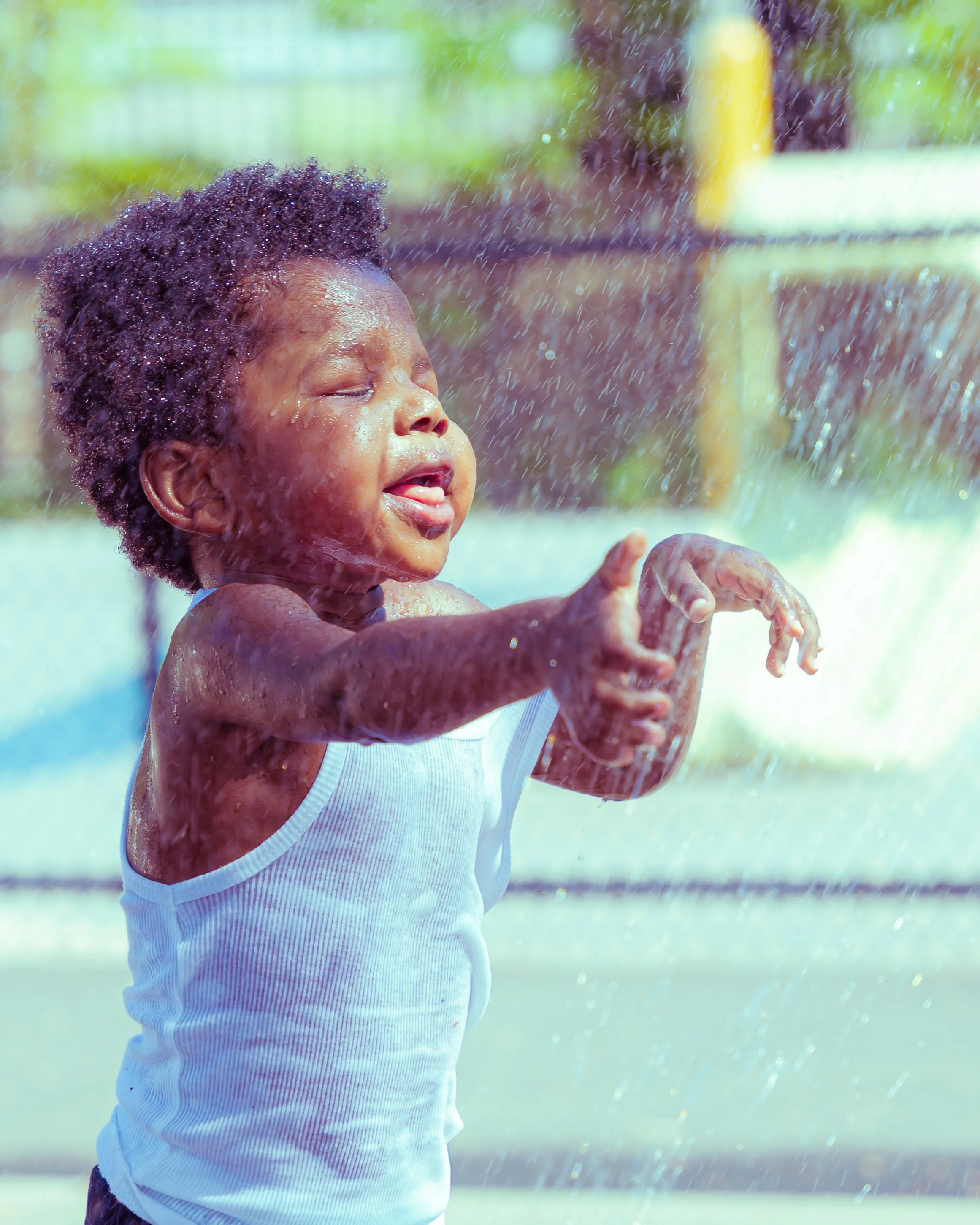 A young child with curly hair wearing a white tank top, playing in the water with a joyful expression.
