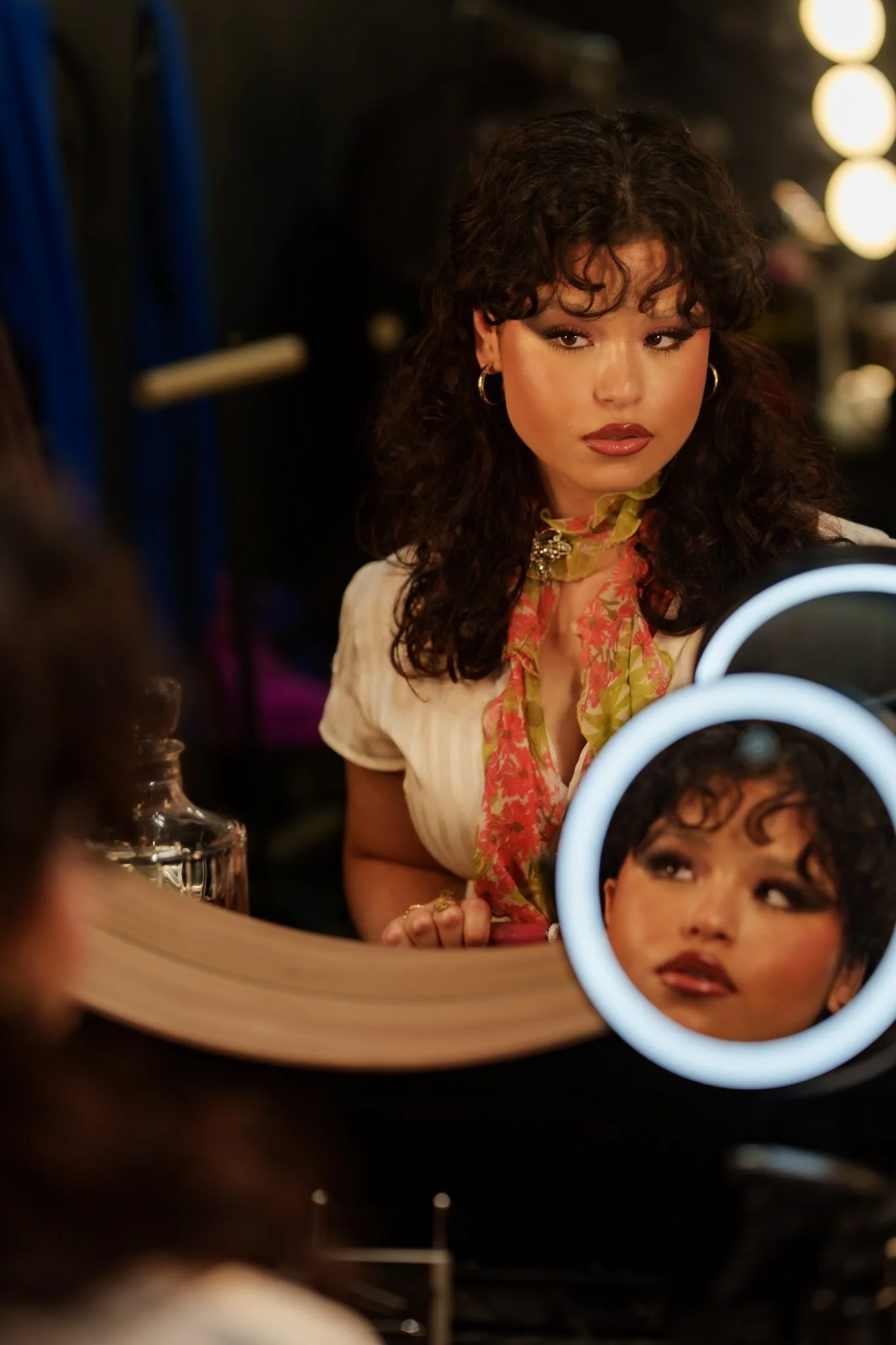 A woman with curly dark hair and makeup looking into a mirror with a illuminated ring light reflection, revealing her face from the reflection.