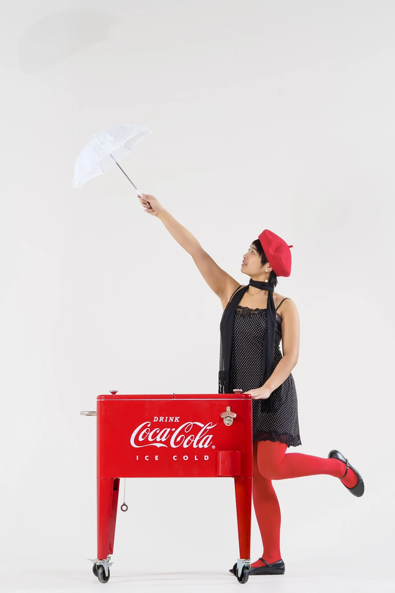 A woman dressed in black with red tights, a red beret, and a black scarf stands behind a red Coca-Cola ice cooler. She is holding a white umbrella high in the air with one hand and balancing on one leg.