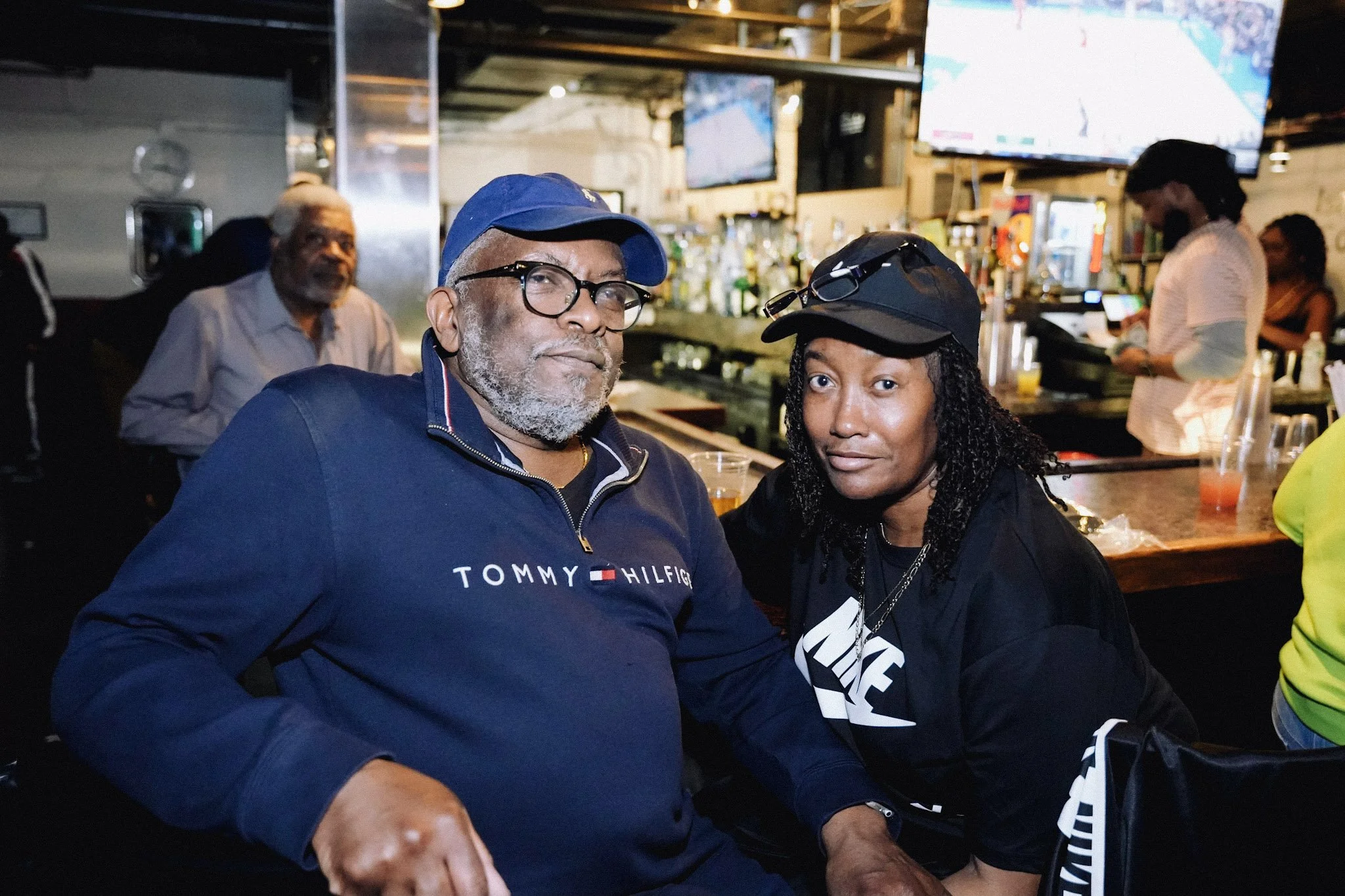 A man and woman sitting together at a bar, both wearing dark clothing and caps, with the man wearing eyeglasses and a Tommy Hilfiger sweatshirt. In the background, there are bar shelving with bottles and a digital screen showing sports.