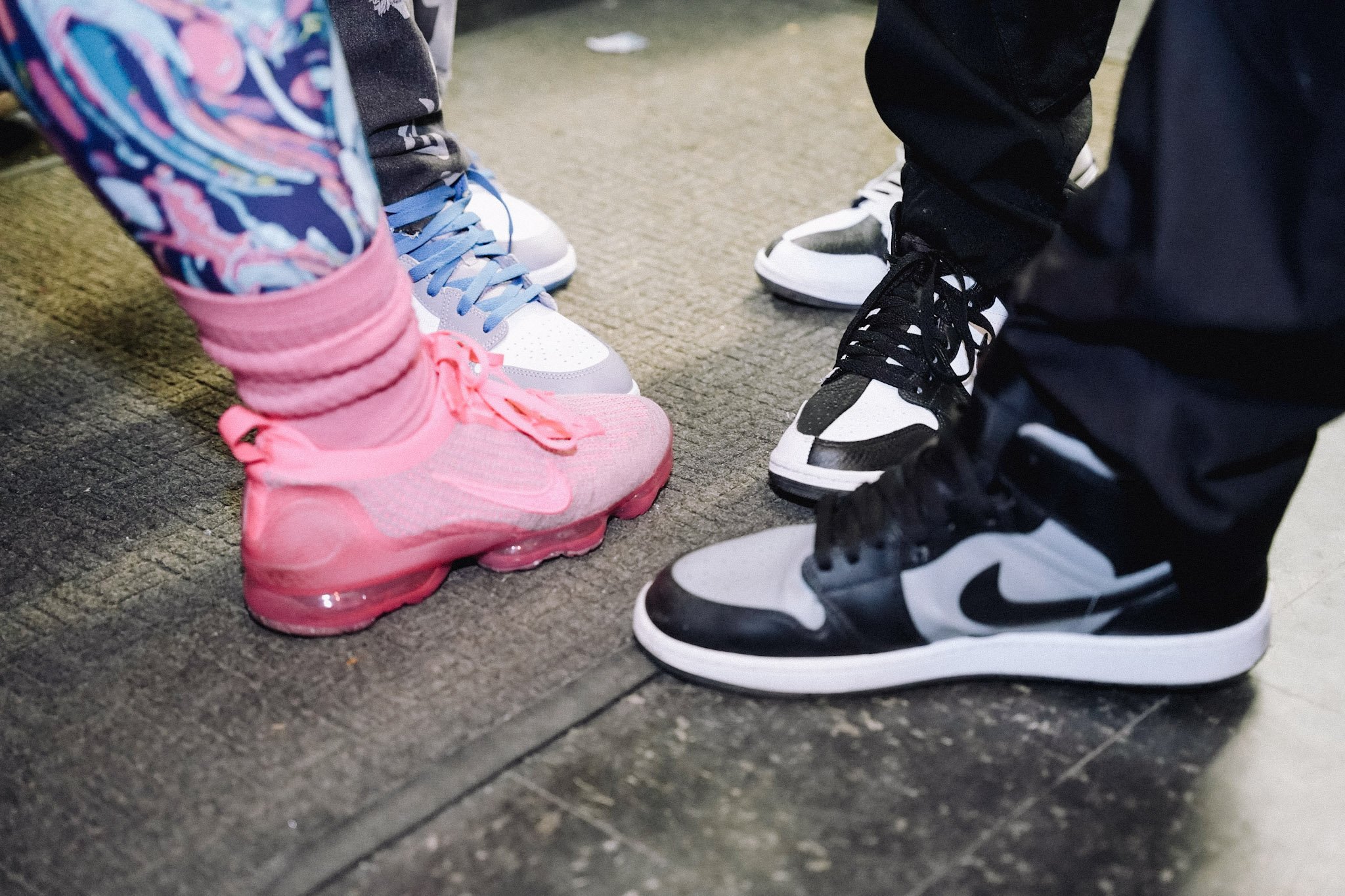 Close-up of three pairs of sneakers on a textured floor, with a person wearing colorful gym pants visible on the left and another person in dark pants on the right.